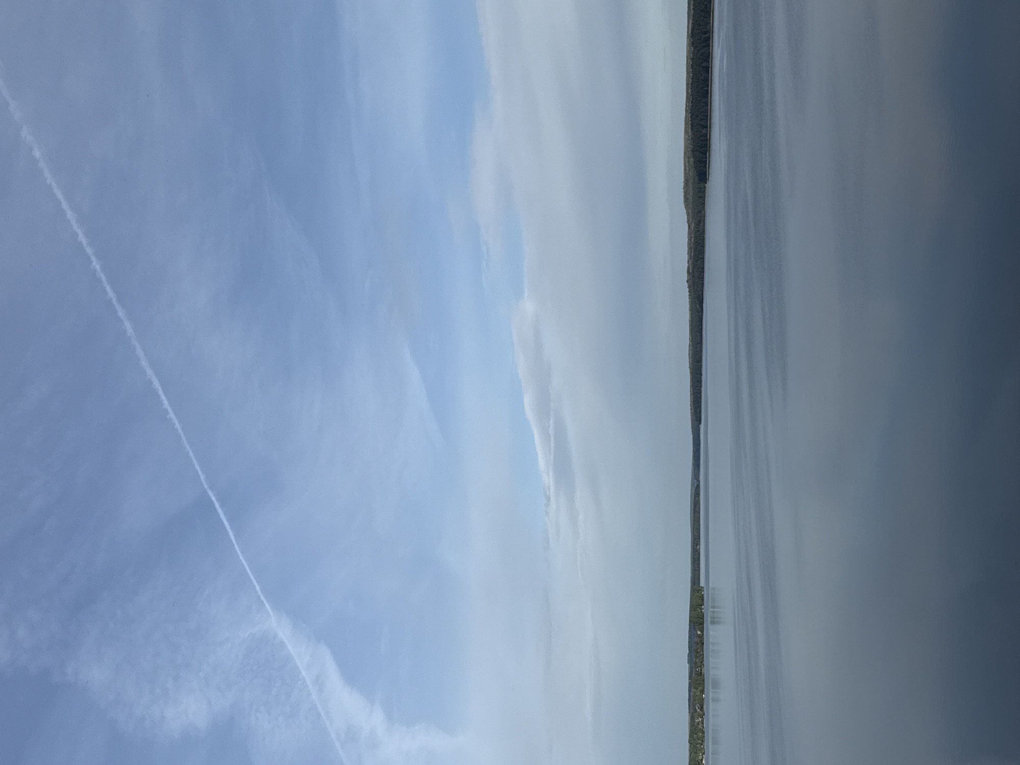 The image depicts a view of Votkinsk Pond in the city of Votkinsk, Russia. The sky is mostly blue with some white clouds, including a prominent contrail from an airplane. The water of the pond occupies the right side of the image and appears calm and reflective. The shoreline is visible as a narrow strip of green vegetation and trees. The image is oriented vertically.
