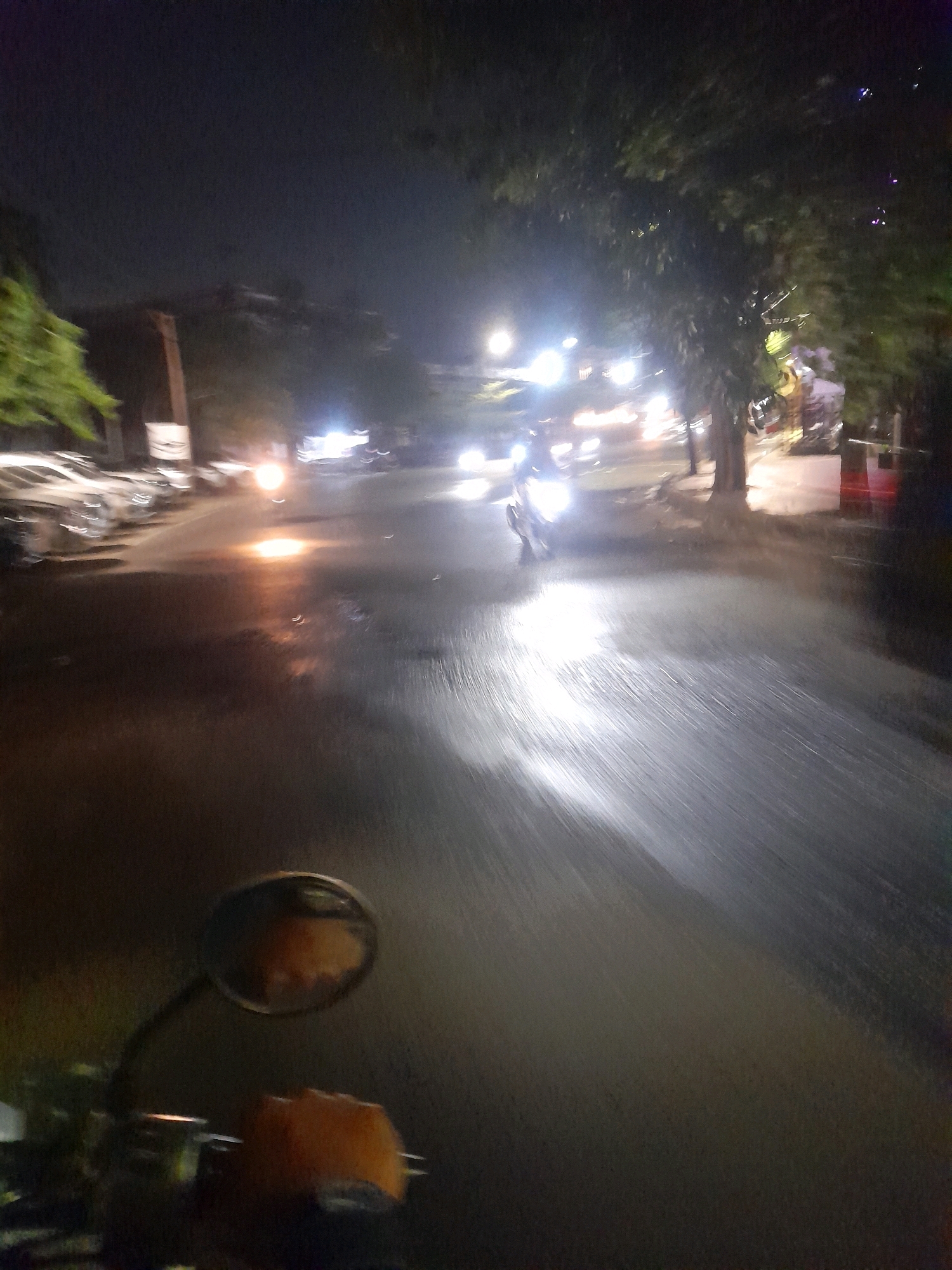 A first-person perspective captures a nighttime scene on a wet city street, likely in Surabaya, Indonesia. The road surface is reflective, showing streaks of light from passing vehicles and streetlights. The image shows a motorcycle's side mirror in the foreground, reflecting a blurred orange and yellow object, possibly the rider's hand or something attached to the handlebars. In the mid-ground, vehicles with illuminated headlights are moving along the street, creating bright streaks of light. Trees with dense foliage line the right side of the street, illuminated by the ambient light. Buildings and parked cars are visible on the left side of the road. The overall impression is one of motion and low light conditions, with the wet pavement contributing to the reflections and the sense of a humid or rainy night.