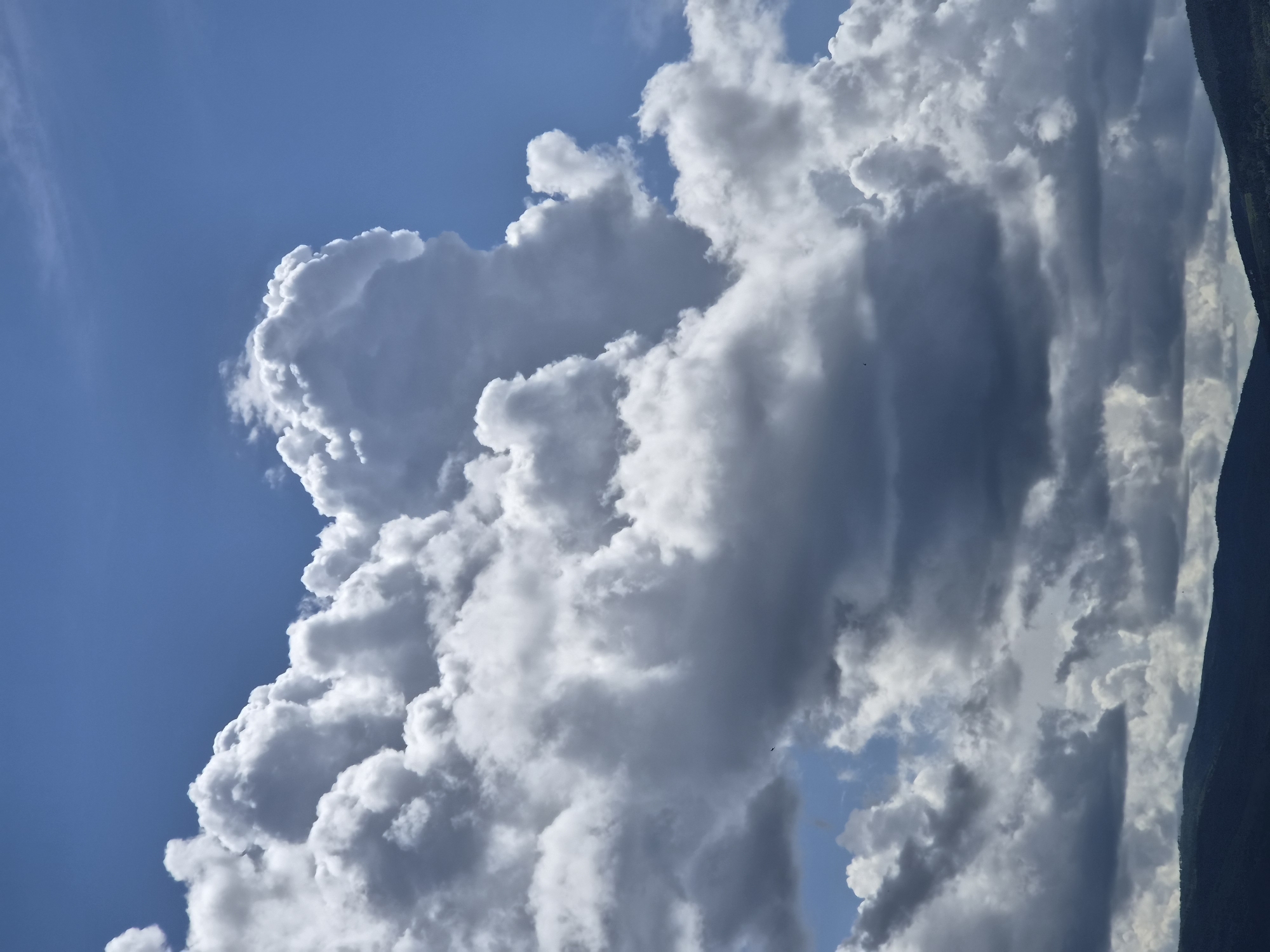 The image displays a dramatic sky with a prominent cluster of large, white, and gray cumulus clouds dominating the center. The clouds are illuminated from above, casting shadows and creating a sense of depth and volume. The sky is a vibrant blue, with lighter patches of blue visible through the cloud formations. In the distance, a dark, undulating silhouette of hills or mountains forms the lower edge of the image. The scene is entirely natural, with no discernible human presence or urban elements. The image is a vertical landscape shot, focusing on the atmospheric conditions above the terrain.