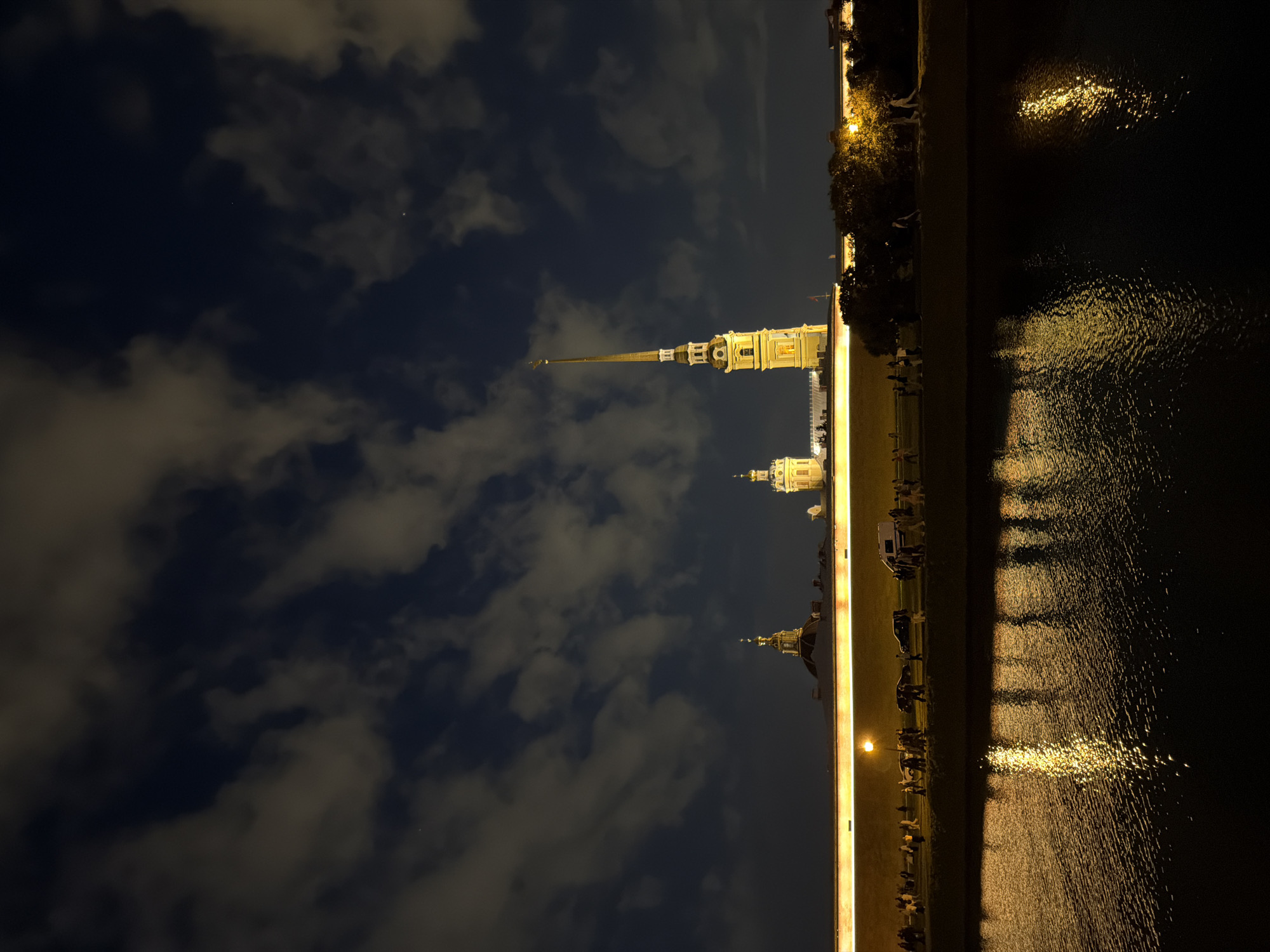 The image depicts the Peter and Paul Fortress in Saint Petersburg, Russia, at night. The fortress walls are illuminated, casting a warm glow, and several domed buildings and a tall spire rise above them. A river or canal reflects the lights of the fortress and the sky, creating shimmering patterns on the dark water. A line of people is visible walking along the embankment in front of the fortress wall, and several cars are parked or moving nearby. The sky is dark with scattered clouds, illuminated by the city lights.