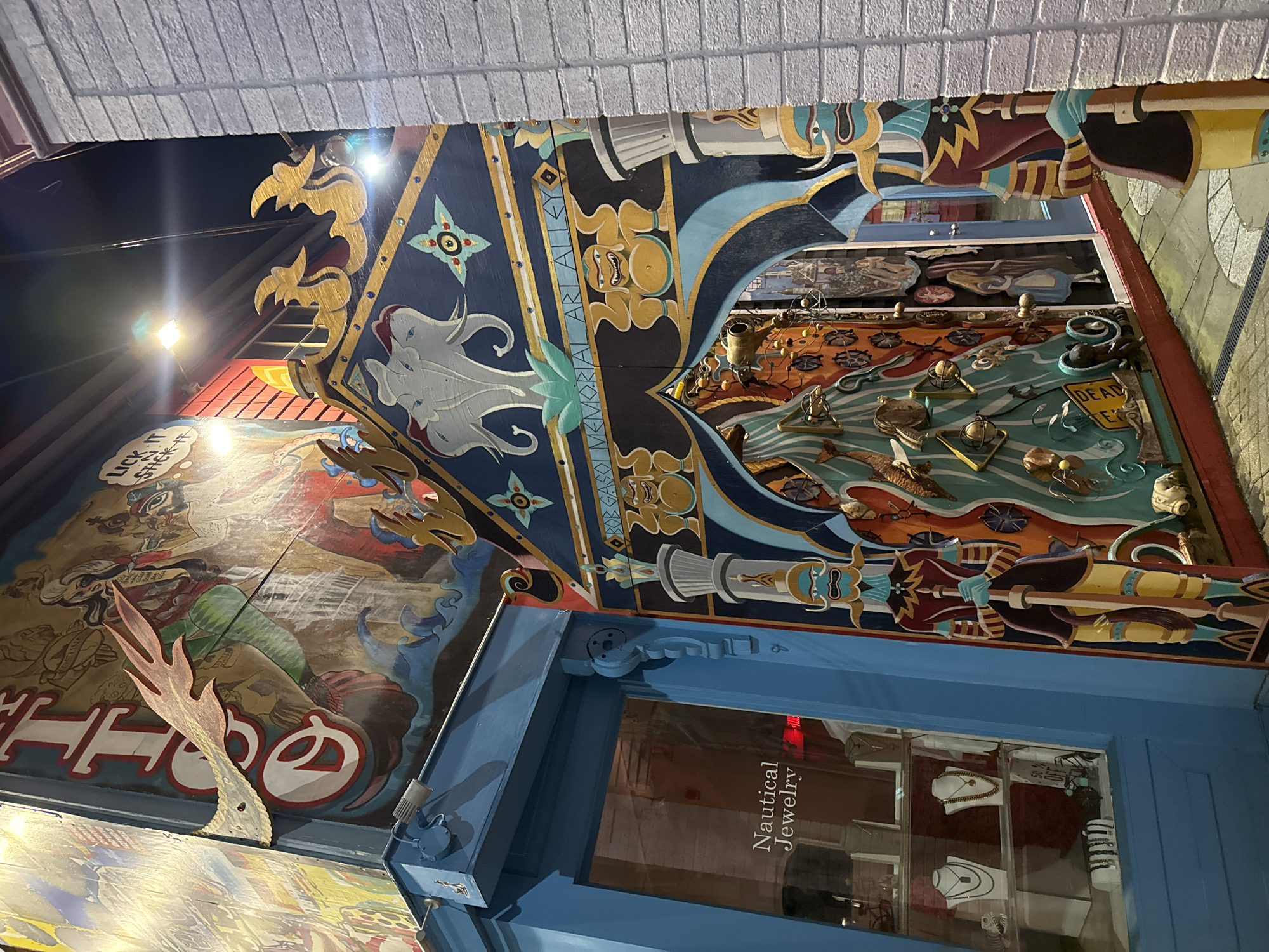 This appears to be a photograph of a section of storefronts in Provincetown, Massachusetts, in the United States. The scene is captured at night, illuminated by street lights, and features a collection of unique and quirky mural artwork.
Visible details include:
* Murals: Distinctive nautical-themed murals, with images of boats, sea creatures, and people. The artwork includes painted figures, carved elements, and various nautical objects arranged in a collage-like fashion.
* Storefronts: The architecture is a mix of brick and painted surfaces, with some buildings having signs for shops. A window reflects an interior of jewelry.
* Text: Sections of text appear within the artwork, including phrases like "BOB GASSO MEMORIAL ART ALLEY" and a shop with a sign reading "Nautical Jewelry."
The overall impression is of an eclectic and artistic atmosphere in Provincetown.
