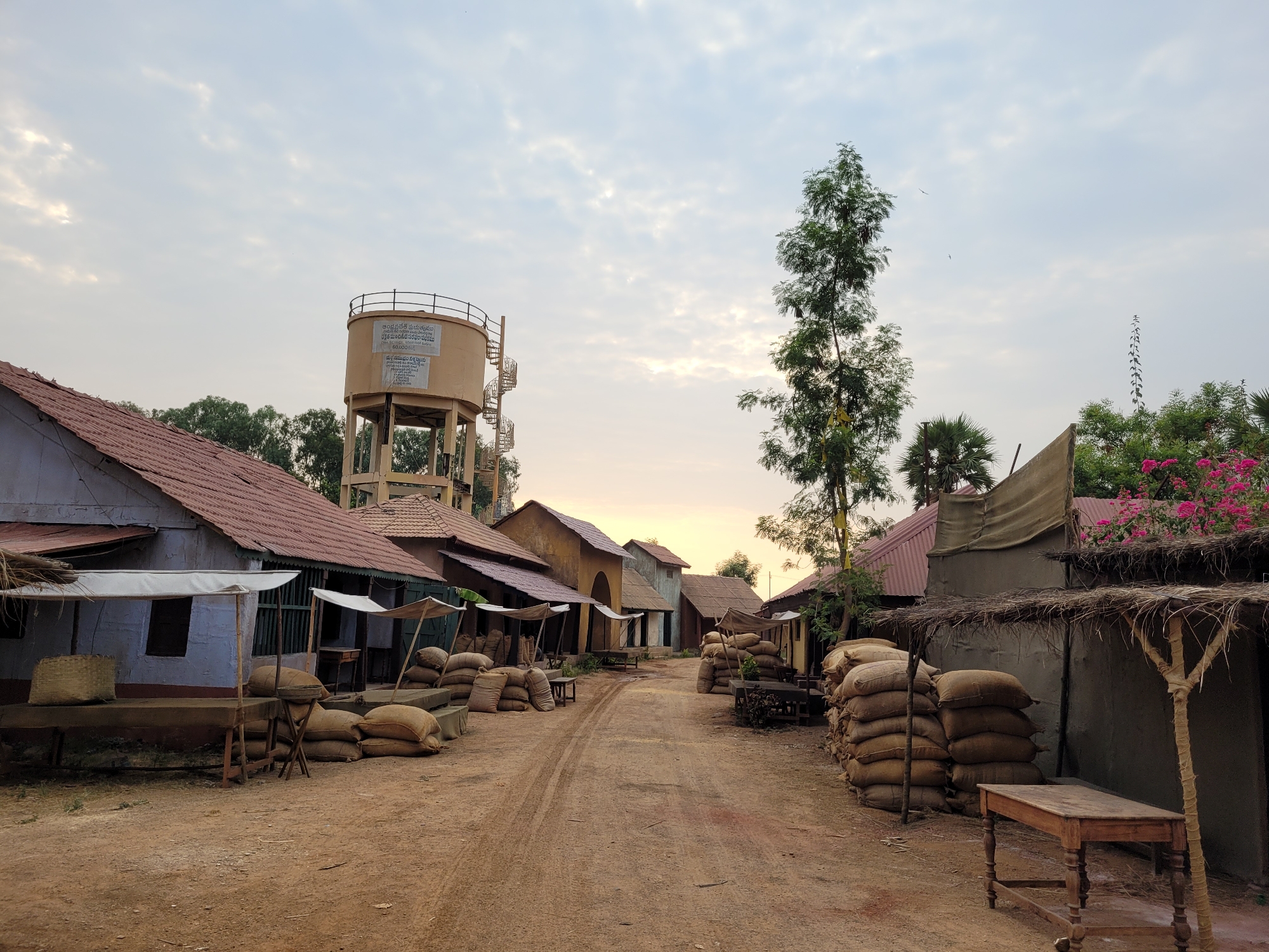 The image depicts a street scene in an unknown location. The street is unpaved and dirt-covered, with visible tire tracks. On either side of the street are low-rise buildings with tiled roofs, some appearing to be market stalls with canvas awnings. Many burlap sacks are stacked along the sides of the street, possibly containing goods for trade. In the background, a water tower with a spiral staircase is visible. The sky is overcast with a hint of light on the horizon. A tall, leafy tree stands towards the right side of the image. There are no people visible in the image.
