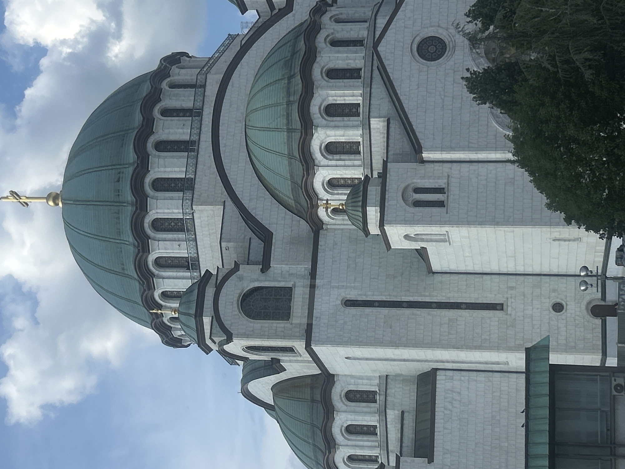 The image shows the Church of Saint Sava in Vracar Urban Municipality, Serbia. The view focuses on the church's dome, topped with a golden cross. The dome is green and ribbed. The church's facade is light grey and made of stone. Arched windows are visible along the dome's base. A tree with dense green foliage partially obscures the church on the right side. The sky is blue with scattered white clouds.