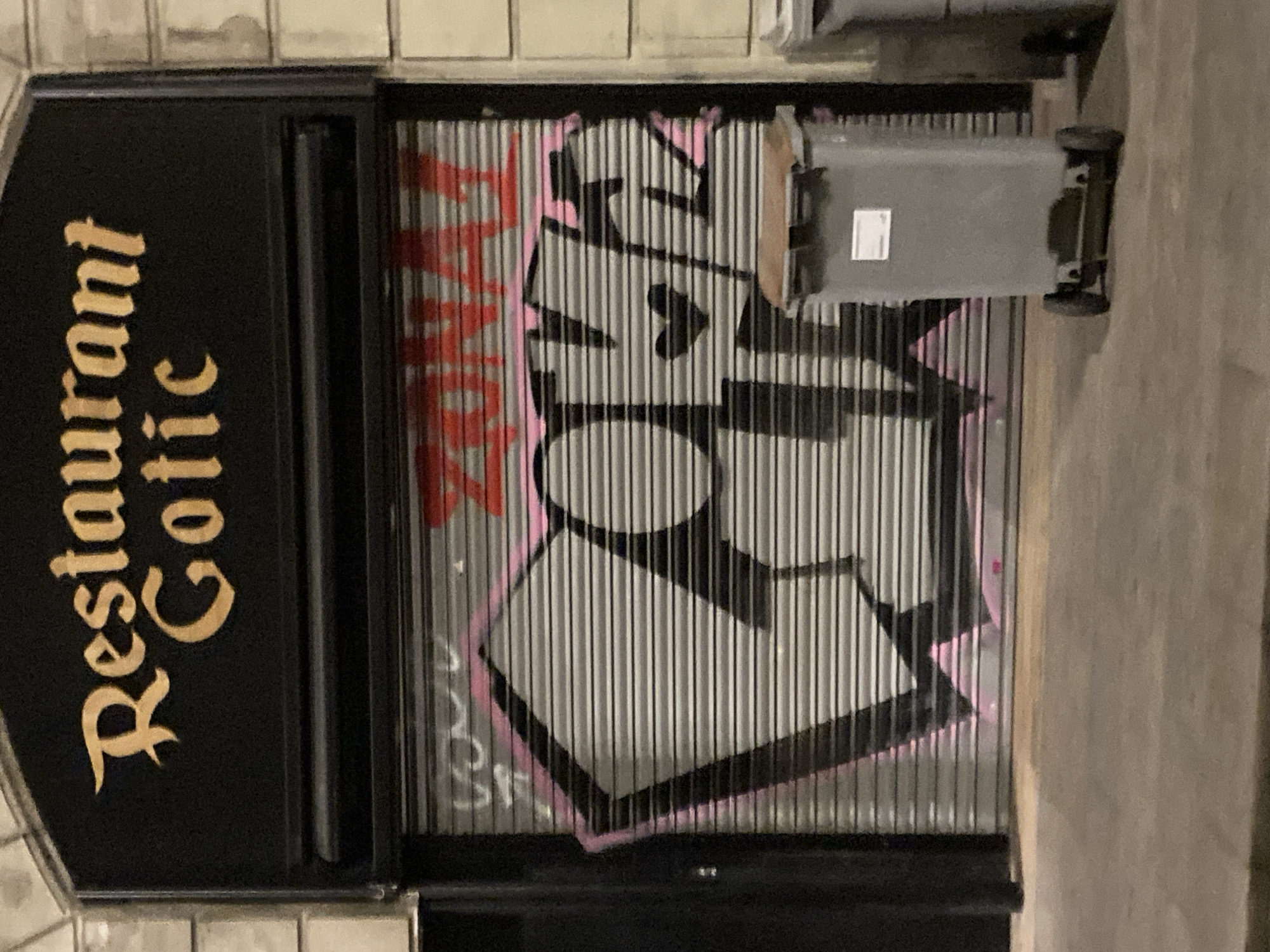 The image captures the facade of a business, likely a restaurant, with a black awning displaying the words "Restaurant Gothic" in gold, ornate lettering. Below the awning, a metal roll-down shutter is partially graffitied. The graffiti includes the word "ZONAL" in red across the top, and a large stylized piece in grey with black outlines and pink accents that appears to spell "LOVE" with a heart shape incorporated. In the foreground, to the right of the shutter, are two wheeled garbage bins, one in focus and grey with a brown lid, and another partially visible bin to its right. The scene is set on a paved street, suggesting an urban environment, possibly in Barcelona, Spain, given the architectural style and the presence of such bins. The lighting suggests it is nighttime or dusk.
