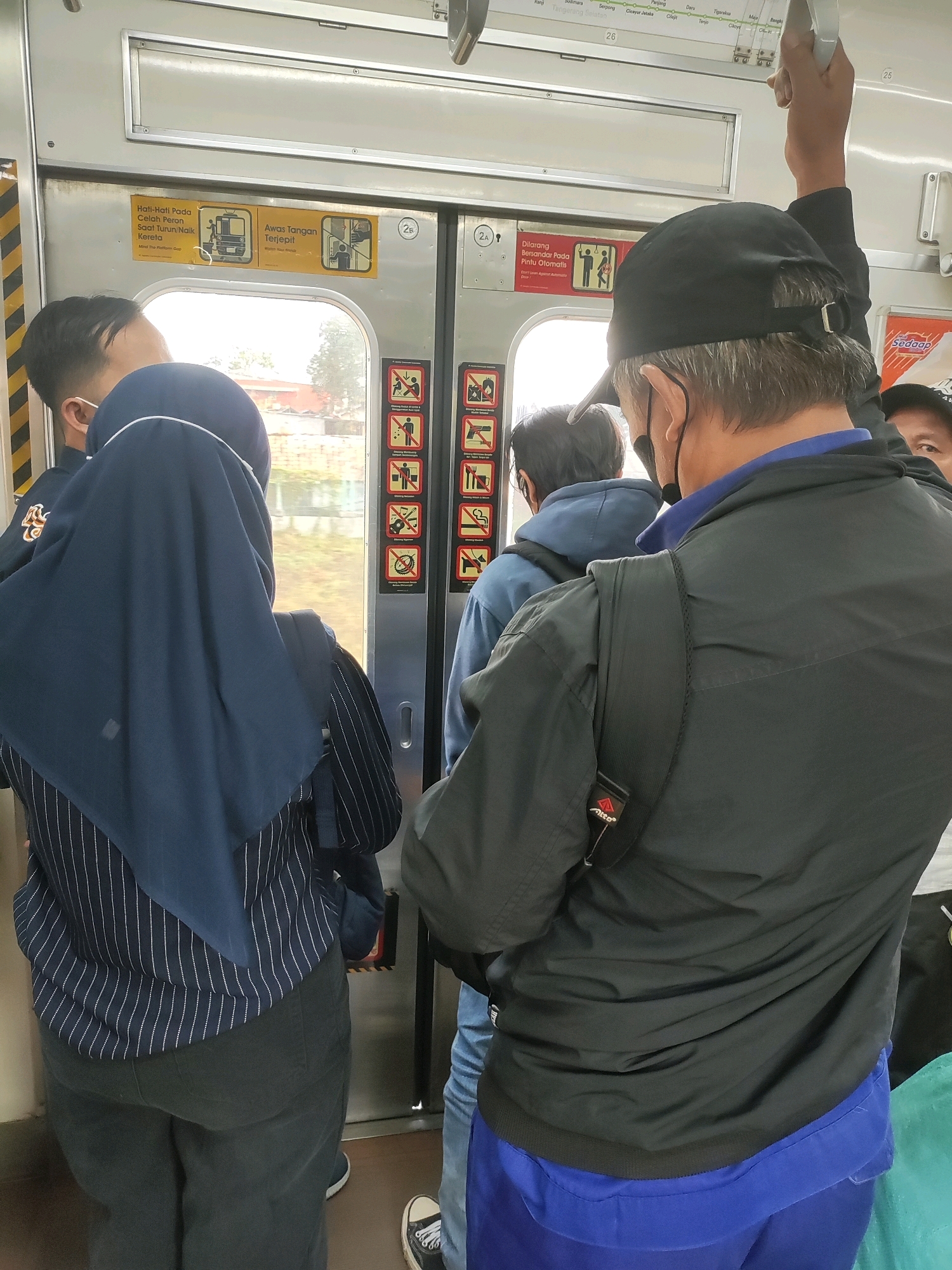 The image shows the interior of a commuter train carriage at Tambun Station, Indonesia. Several passengers are visible. In the foreground, a person wearing a black baseball cap and a black jacket with a blue collar is facing away from the camera, holding onto a handrail. To the left, a person wearing a navy hijab and a striped shirt is also facing away from the camera. To the right and further back, another person in a blue hoodie is visible. On the train door, there are several yellow and red safety warning signs. The environment appears to be an indoor train compartment with windows showing greenery and buildings outside.