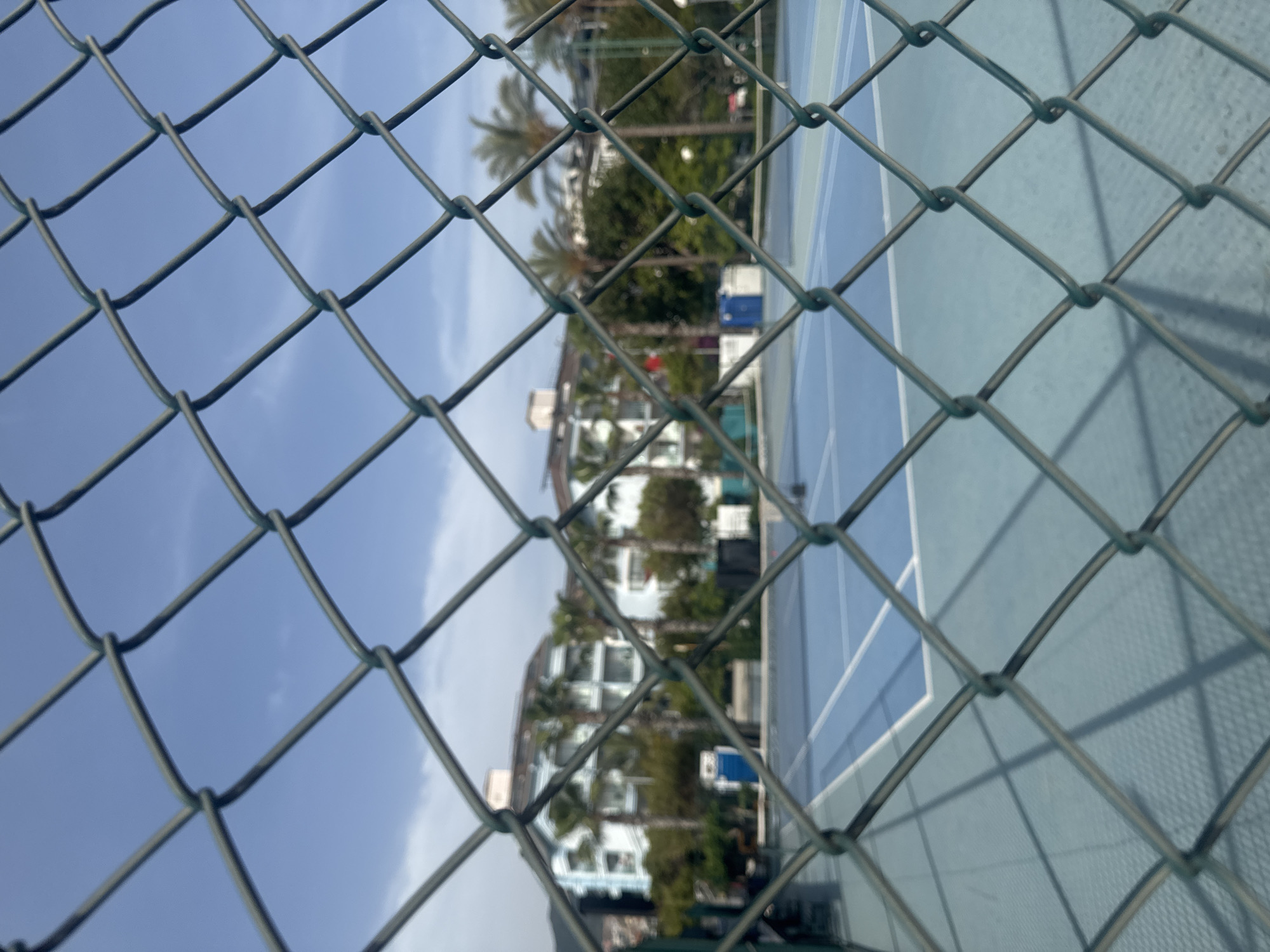 The image depicts a tennis court, view obstructed by a chain-link fence. Beyond the fence, palm trees and a multi-story building are visible. The sky appears blue with some cloud cover. This scene is consistent with a location in Alanya, Turkey, based on the architecture and vegetation.