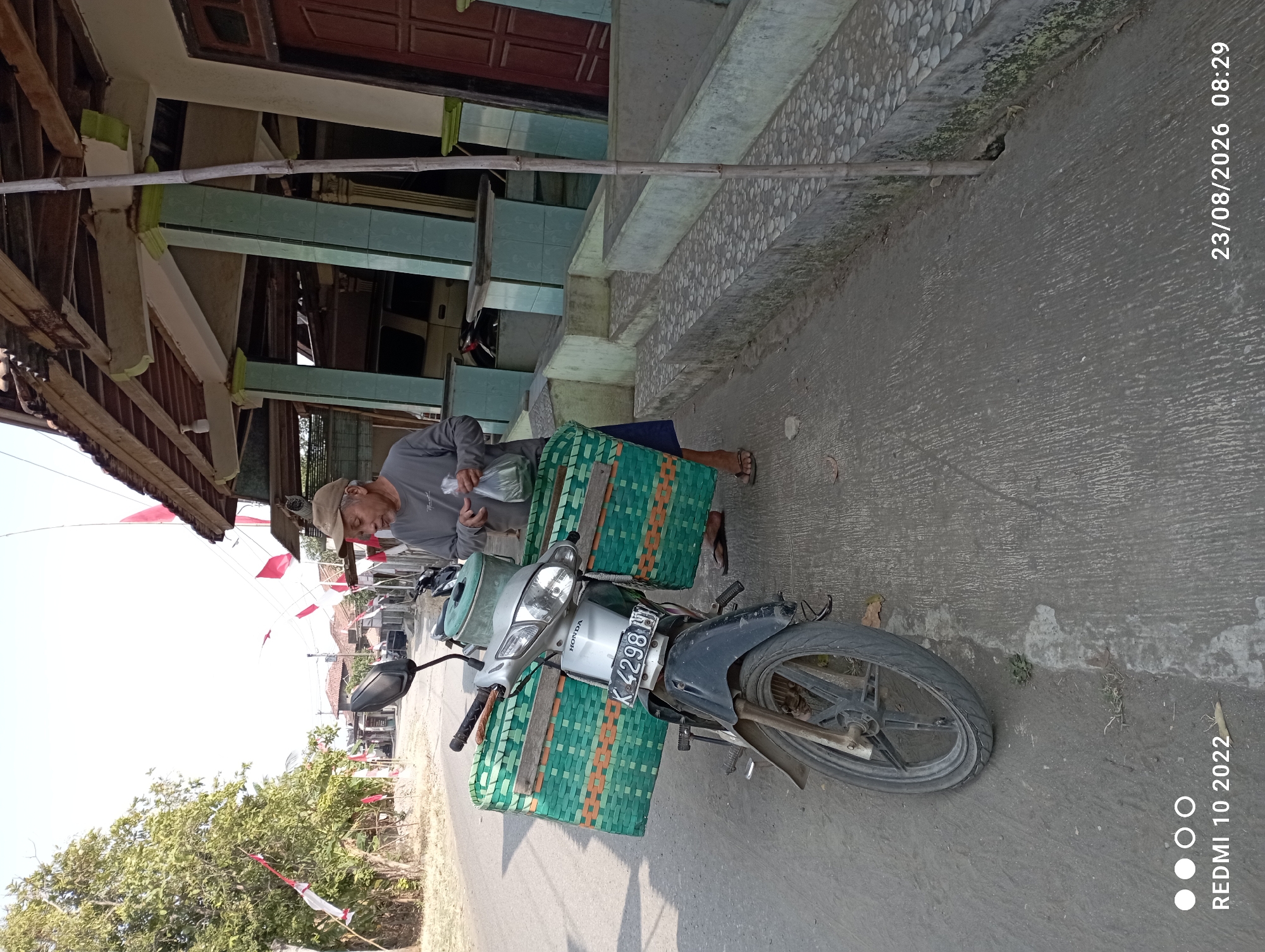 In Dukuhseti, Indonesia, an elderly man stands next to a motorcycle parked on a street, preparing to load or unload bags. The motorcycle is equipped with two large green woven baskets on either side, suggesting it's used for transport or sales. The man, wearing a cap and a long-sleeved shirt, is holding plastic bags. The background shows a house with a traditional Indonesian architectural style, featuring an open veranda with pillars. Indonesian flags are visible, adding a festive or commemorative atmosphere. The environment appears to be a rural or semi-rural area, with trees and a clear sky. The timestamp on the image indicates it was taken on August 23, 2026, at 08:29.