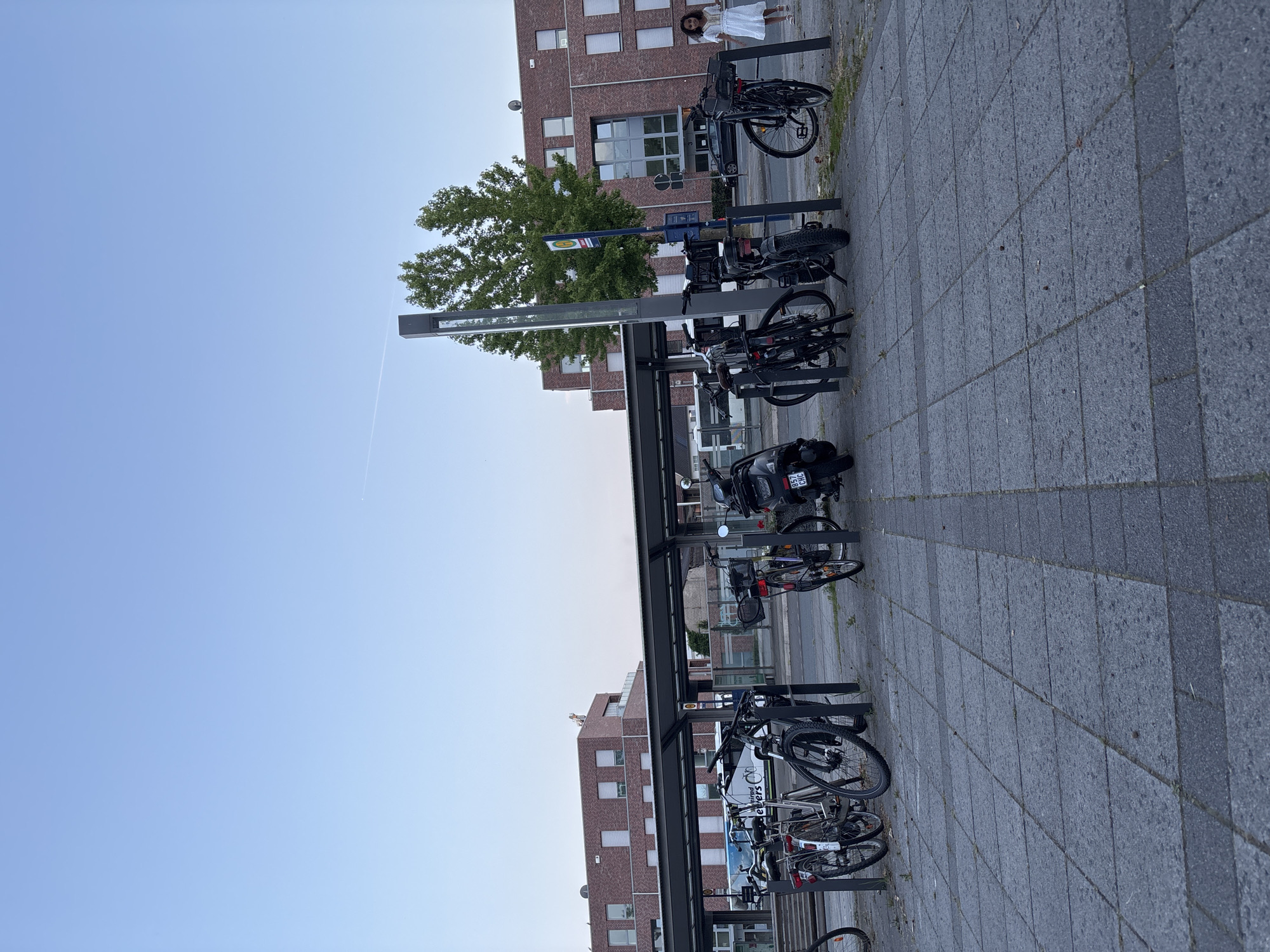 This is a photograph taken at the train station in Gronau, Germany. In the foreground, a paved area with square gray tiles leads to a covered bicycle rack filled with parked bicycles and a scooter. A woman in a white dress walks away from the camera on the right side of the image. Behind the bike rack, a bus is visible, and beyond that, a modern brick building with multiple windows stands under a clear blue sky with a faint contrail. The overall scene depicts an urban environment at what appears to be late afternoon or early evening, given the soft lighting.
