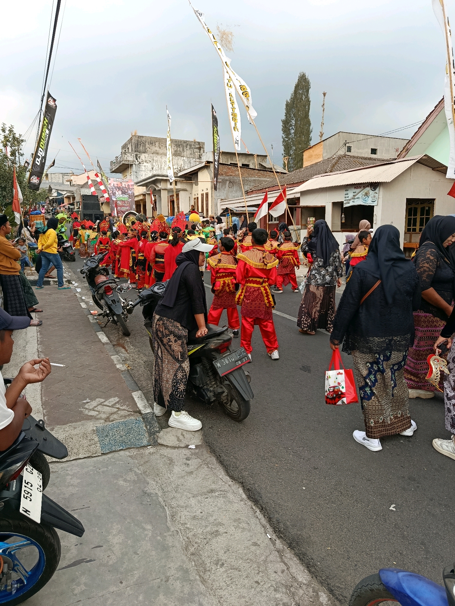In Gubukklakah, Indonesia, a street event is underway. A parade is proceeding down a paved road lined with buildings and trees. Many participants are dressed in traditional Indonesian attire, featuring red and gold costumes. Several individuals are wearing black headscarves and patterned skirts. Some people are watching the parade from the side of the road, while others are actively participating in the procession. Motorcycles are parked along the street, and flags are displayed, indicating a festive occasion. The sky is overcast, suggesting it might be a cloudy day.