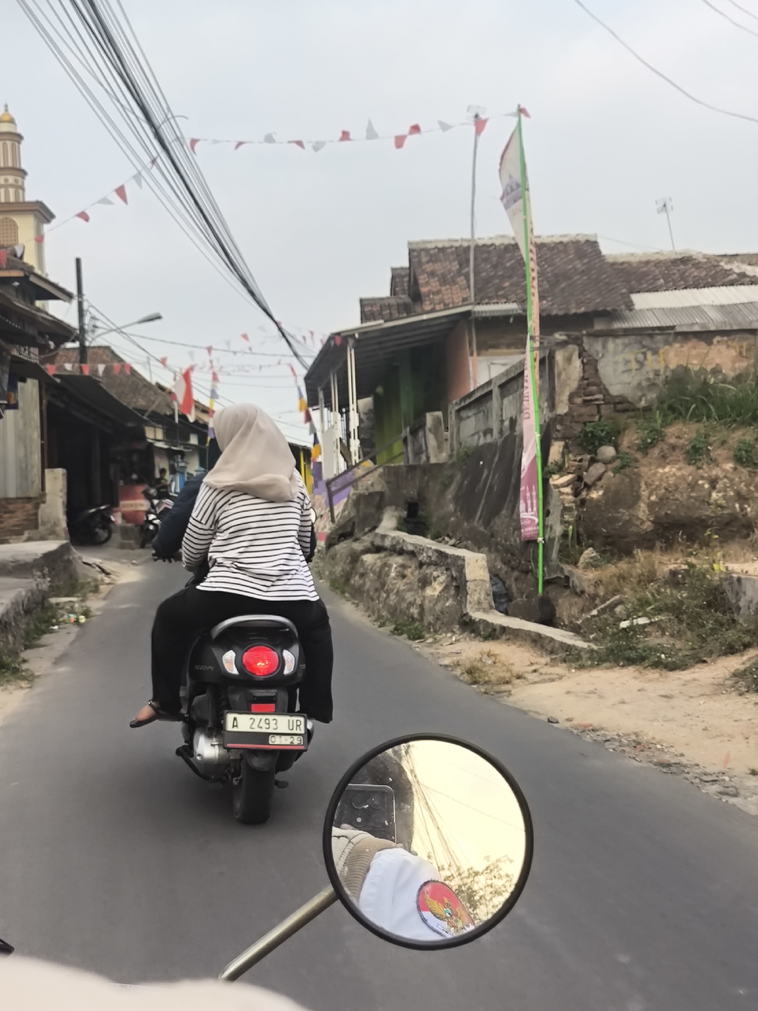 The image depicts a street scene in Cilegon, Indonesia, likely on a cloudy day. A person wearing a hijab and a striped shirt is riding a scooter. The scooter has a license plate reading "A 2493 UR" and "01-29". In the foreground, a rearview mirror reflects the rider's arm, which has an emblem on it, and a blurred background of trees and buildings. Along the street, red and white flags are strung across wires, suggesting a local celebration or holiday. Buildings line both sides of the road, some appearing to be shops or homes, with a mosque visible in the distance. The road is paved, and a ditch runs alongside it on the right. The environment appears to be a mix of urban and semi-rural elements.