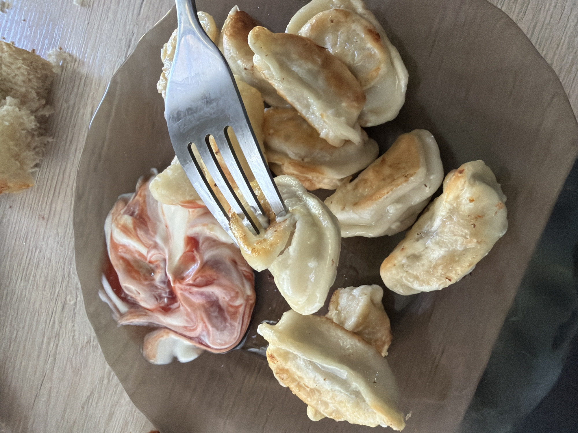 The image shows a close-up overhead view of a plate of dumplings on a wooden surface. A fork is piercing one of the dumplings, and a swirl of pink and white sauce is next to it. Some pieces of bread are visible to the left of the plate. The dumplings appear to be pan-fried, with some browned edges. The sauce is likely a mix of mayonnaise and ketchup or another red sauce. The scene suggests a meal being prepared or eaten. There are no people visible, and the time of day and weather are not indicated. The location context is provided as Shumki, Russia, but there are no specific visual cues in the image to confirm this. The lighting suggests an indoor setting with artificial light.
