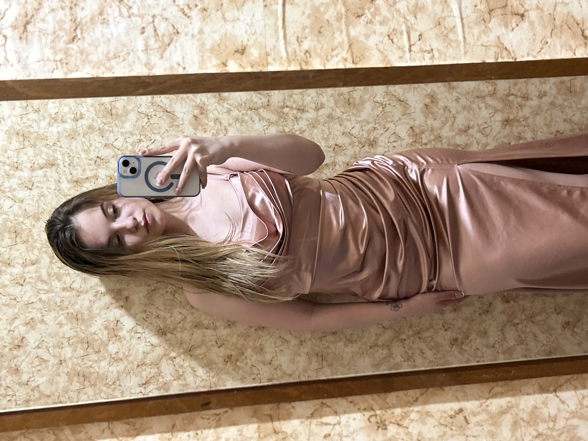 A woman is taking a selfie in a mirror. She is wearing a pink dress. The background appears to be a wall with a marble-like pattern and wooden framing. The location is unknown; no evidence indicates it is in Plovdiv, Bulgaria.