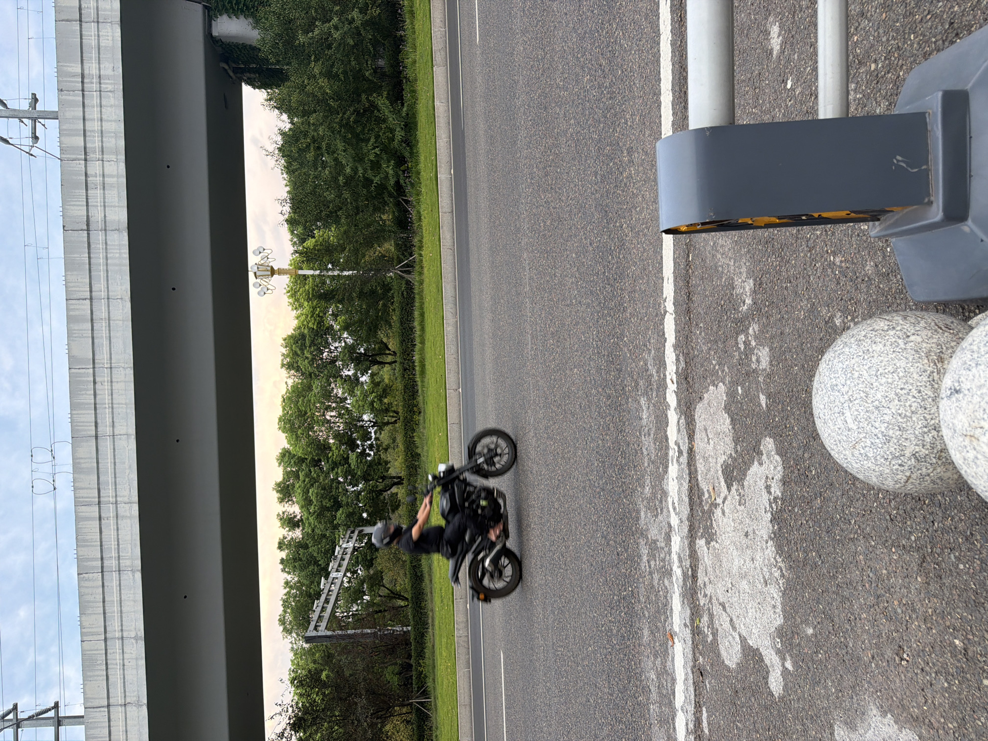 The image depicts a street scene in Jiangjin, China. A person wearing a black helmet and black clothing is riding a black motorcycle in the left lane of a paved road. The motorcycle is in motion, evidenced by the slight blur. The road is bordered by a grassy median and then a dense line of green trees. Above the trees, a large concrete overpass is visible, supported by columns. Power lines and catenary wires are strung above the overpass. In the foreground, the edge of the road is visible, with some white markings and patches of lighter colored pavement. Two large, round, speckled granite spheres are positioned in the lower foreground, along with a dark gray traffic barrier with yellow reflectors. The sky is overcast with hints of sunset colors.