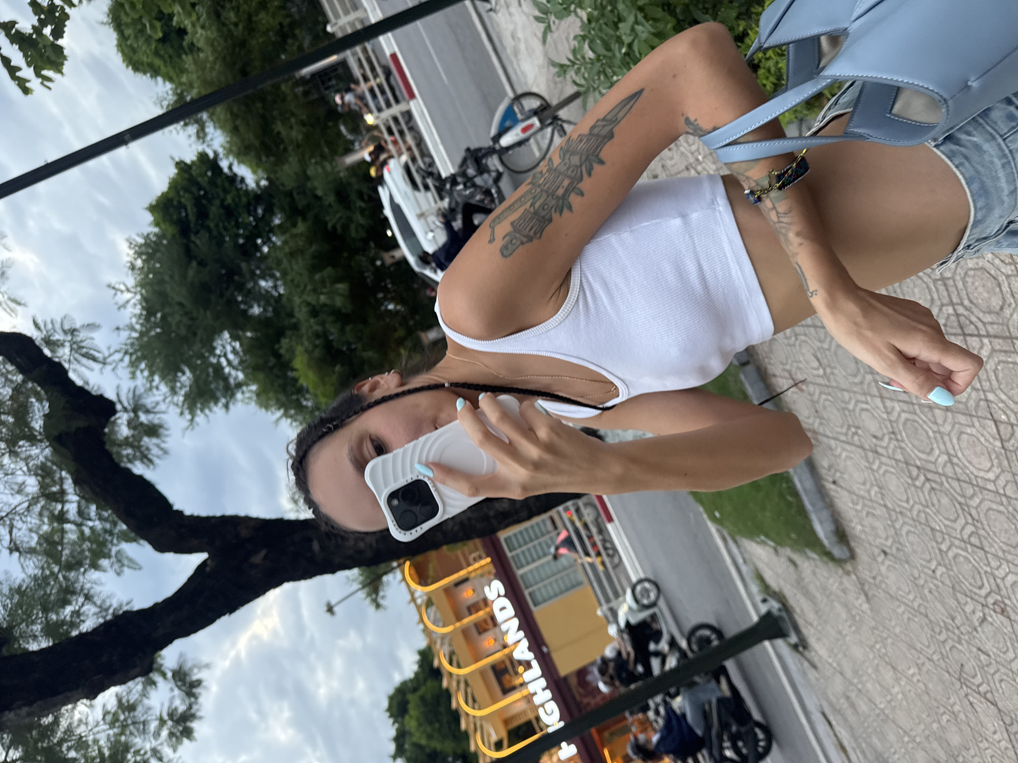 A young woman with dark hair in braids is taking a selfie with her white phone case in the foreground, holding it up to her face. She is wearing a white cropped tank top and light blue denim shorts. She has several tattoos on her left arm, including what appears to be a dagger design. She is also carrying a light blue handbag. In the background, a building with illuminated signage that reads "HIGHLANDS" is visible. The scene is set on a paved sidewalk next to a street where several motorbikes and a white car are present, indicating an urban environment in Hà Nội, Vietnam. The sky is overcast.