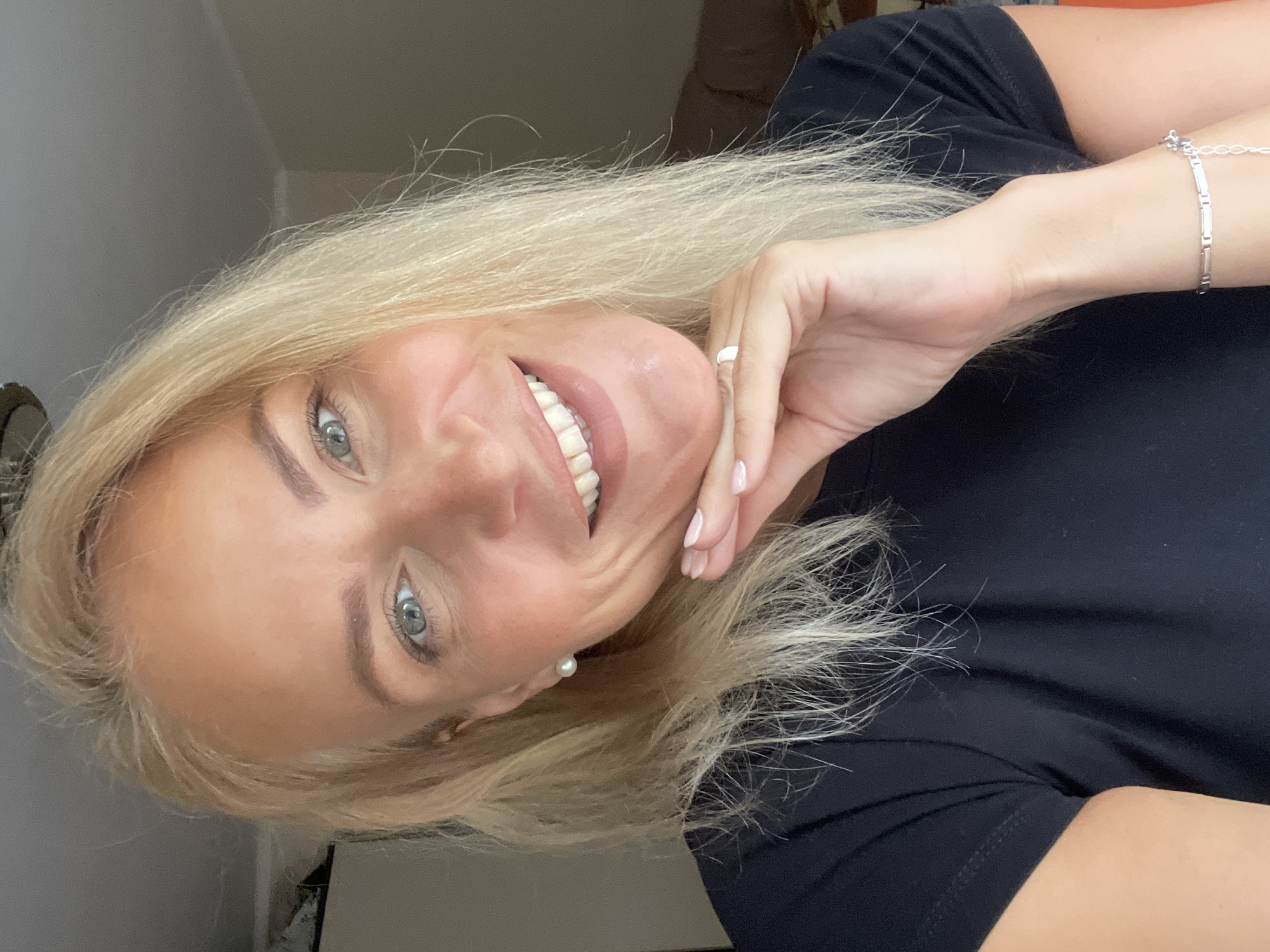 A woman with blonde hair and blue eyes is smiling at the camera. She is wearing a black shirt, a silver bracelet, and a small pearl earring. Her right hand is touching her chin. The image appears to be taken indoors. No information is present to connect the image with Sochi, Russia.