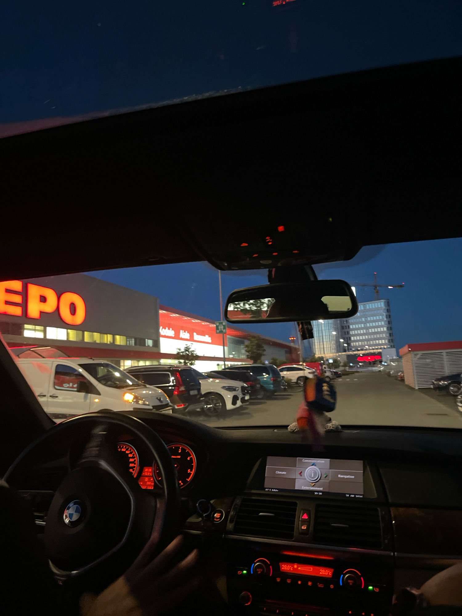 The image was taken from inside a BMW vehicle, looking forward through the windshield at a parking lot and a commercial building at dusk. The commercial building has large, illuminated orange letters spelling out "EPO". To the right of the building, there are red accents and a smaller, red-roofed structure. In the distance, a tall, modern glass building is visible, along with construction cranes.
The parking lot contains several cars, including white vans, black SUVs, and white sedans. The lighting suggests it is early evening. The car's dashboard is visible in the foreground, with illuminated gauges and a climate control display showing 28.0°C. A hand is visible on the steering wheel.
Based on the building signage ("EPO") and the modern architecture, the location is likely Tallinn, Estonia, specifically a commercial area. The overall scene depicts a person in their car, parked outside a shopping center or business district in the city at twilight.