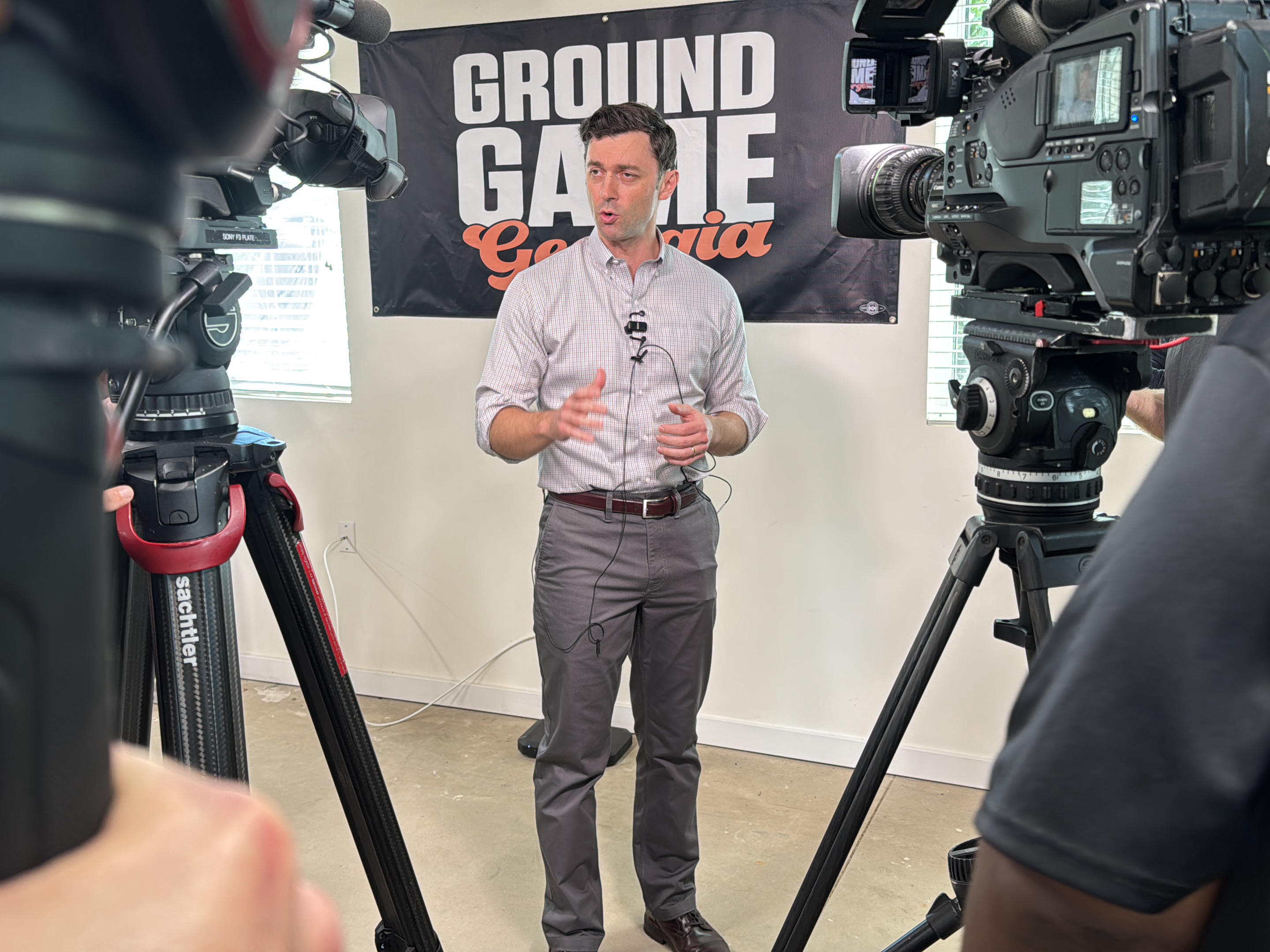 The image shows a man standing in front of a "Ground Game Georgia" banner, likely in Decatur, United States, based on the banner's text. He is wearing a light-colored, button-down shirt, grey pants, a brown belt, and brown shoes. A lavalier microphone is clipped to his shirt. He is positioned between two professional video cameras mounted on tripods. The person on the right side of the image is wearing a dark gray shirt. The background includes a white wall with an electrical outlet and a window. The banner is black with white and orange lettering. The floor appears to be light concrete.
