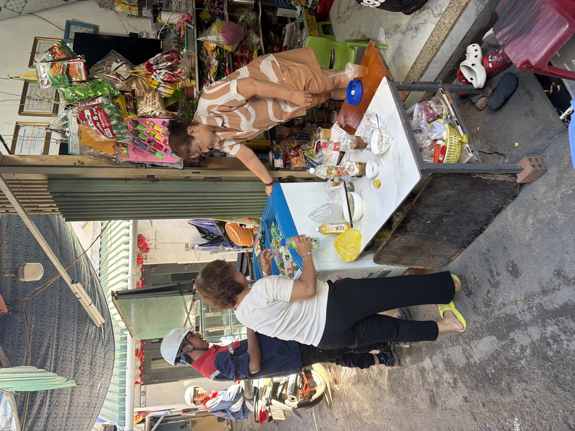 The image depicts a street scene in Bac Nha Trang Ward, Vietnam. In the foreground, a woman in a patterned short-sleeved shirt and shorts is interacting with a refrigerator, holding a bottle. Next to her, a table is laden with drinks and food items in bowls and cups. A person in black pants and a white t-shirt stands behind the table, also near the refrigerator. To the left, a man wearing a blue and red shirt and a white helmet is standing next to a motorcycle. Further left, another person in a blue jacket and white helmet is seated on the motorcycle. The environment appears to be an outdoor market or a shopfront with shelves filled with various goods, including packaged snacks, drinks, and other items. The ground is paved with rough concrete, and there is overhead covering made of fabric and corrugated metal.