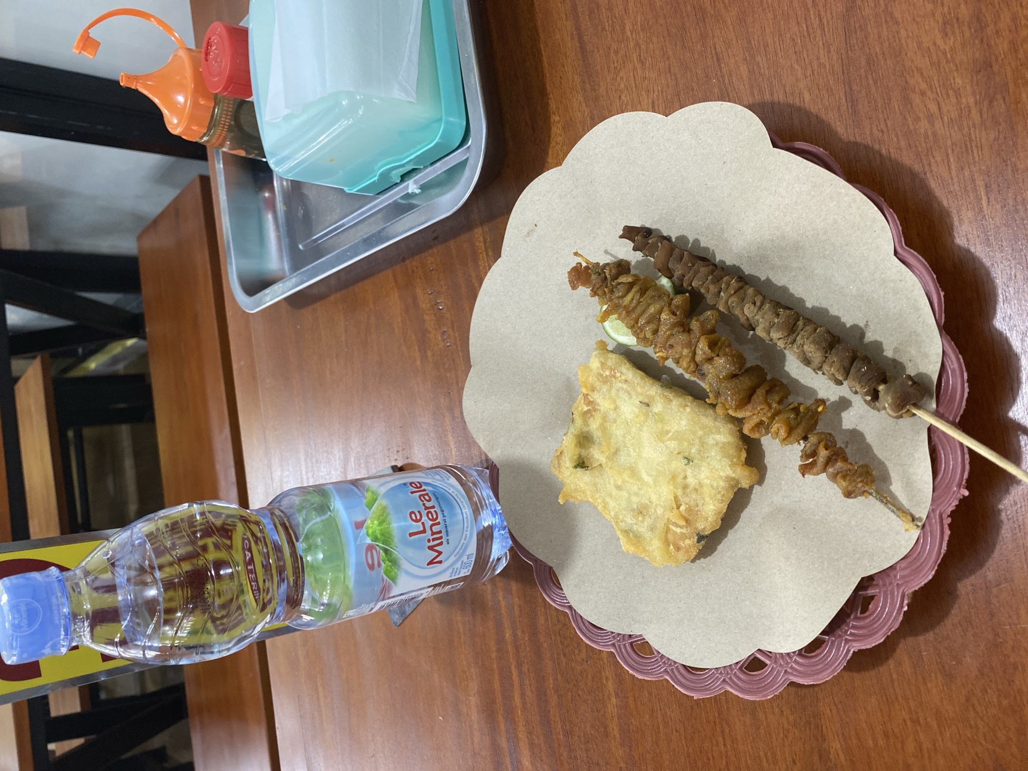 A bottle of Le Minerale bottled water sits on a wooden table next to a pink plate with a scalloped edge. On the plate, a piece of fried tempeh and two skewers of grilled meat are placed on brown paper. In the background, a metal tray holds an orange sauce dispenser and a light blue container, likely containing tissues. The setting appears to be a casual dining establishment, possibly a warung or restaurant, in North Jakarta, Indonesia. The lighting suggests it is daytime.