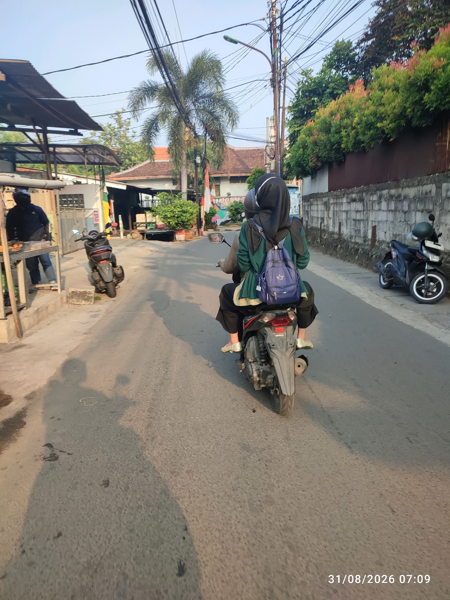 The image captures a street scene in East Jakarta, Indonesia, at 07:09 on August 31, 2026. In the foreground, a person wearing a hijab and a green jacket is riding as a passenger on a scooter, their back to the camera. They are carrying a blue backpack adorned with a white star. The scooter's license plate is partially visible and appears to be for a vehicle registered in Indonesia. The rider is wearing black pants and white slip-on shoes. The scooter itself is gray and black.
To the left of the road, a man in a dark shirt and blue jeans stands near a food stall or market stall. He is facing away from the camera and appears to be attending to items on display. The stall has a corrugated metal roof and is supported by wooden poles. A parked motorcycle, dark in color, is visible next to the stall.
Further down the street, another dark-colored motorcycle is parked on the right side of the road, facing away from the viewer. It has a green helmet on the handlebars. The street is paved and appears to be a residential or local commercial area, with houses and buildings visible in the background. Green trees and foliage line the wall on the right side of the street, and a tall palm tree is prominent on the left. Utility wires are strung overhead. The lighting suggests it is early morning.