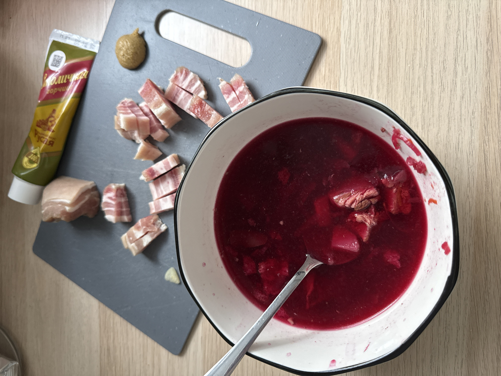 An overhead shot captures a meal or food preparation scene on a light wooden table. A bowl of deep red soup, likely borscht, occupies the right side of the frame, with a spoon resting in it. Visible in the soup are pieces of meat and what appear to be vegetables.
To the left of the bowl, on a grey cutting board, are several cubed pieces of bacon or pork fat (salo), some with skin. A larger, uncut piece of raw pork belly/fat is also visible on the board. A small dollop of yellow-brown mustard is placed directly on the cutting board, ready for consumption. Next to the mustard dollop is a tube of mustard. The text on the tube is in Cyrillic, clearly showing "Столичка" (Stolichka) and "Горчица" (Gorčica, meaning Mustard). The brand name "Руся" (Rusya) is also visible, along with a QR code on the top left of the tube.
The image depicts a typical Eastern European meal, with borscht, salo, and strong mustard as accompaniments, consistent with the given location context of Moscow, Russia. The setting appears to be a casual home environment.