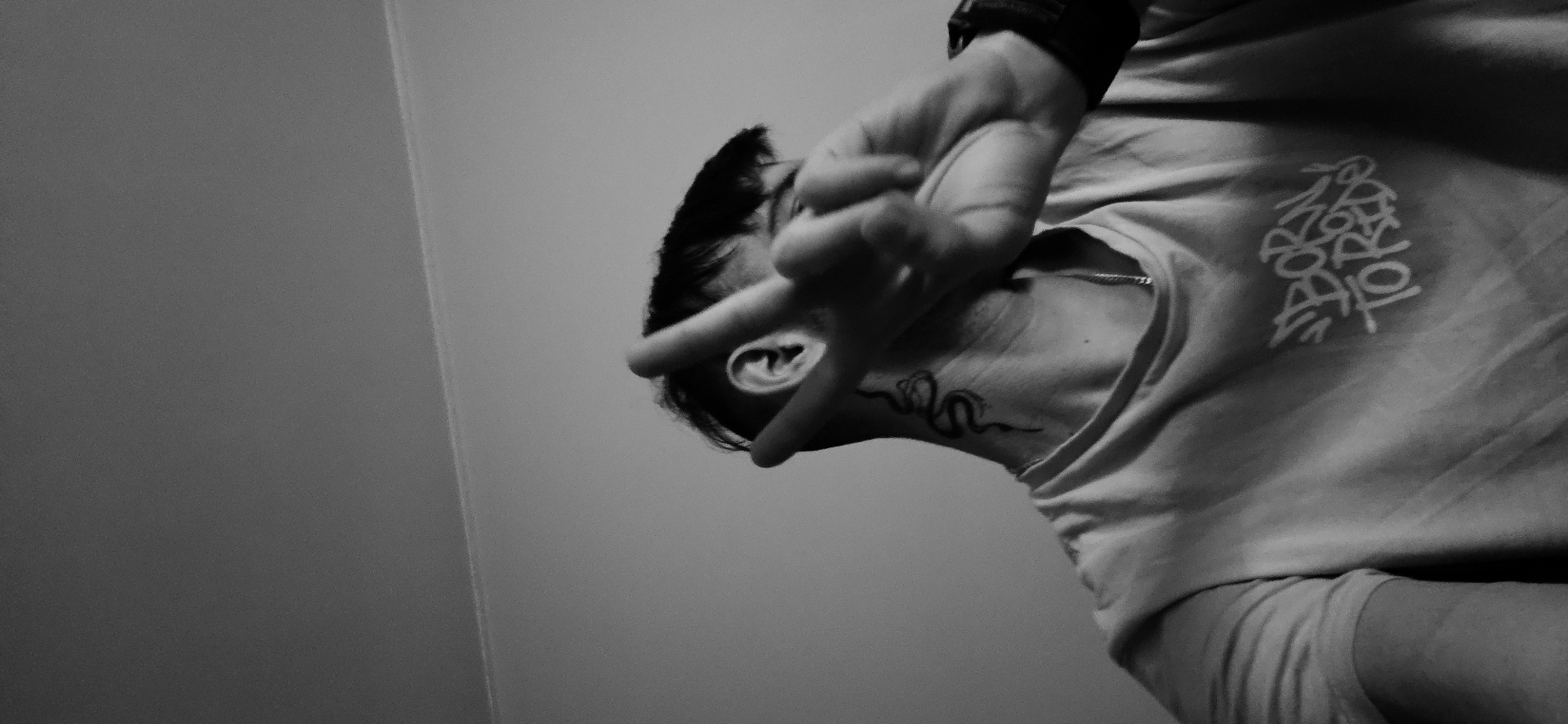 The black and white image shows a person from the chest up, facing the right side of the frame. The person is partially obscuring their face with their hand, which is raised to head level. A wristwatch is visible on their left wrist. The person has short, dark hair and a tattoo of a snake on their neck. They are wearing a light-colored t-shirt with the words "BORN TO TRADE" printed on it. The background is a plain gray wall. There is no information in the image to determine the location in the city of Plovdiv, Bulgaria.
