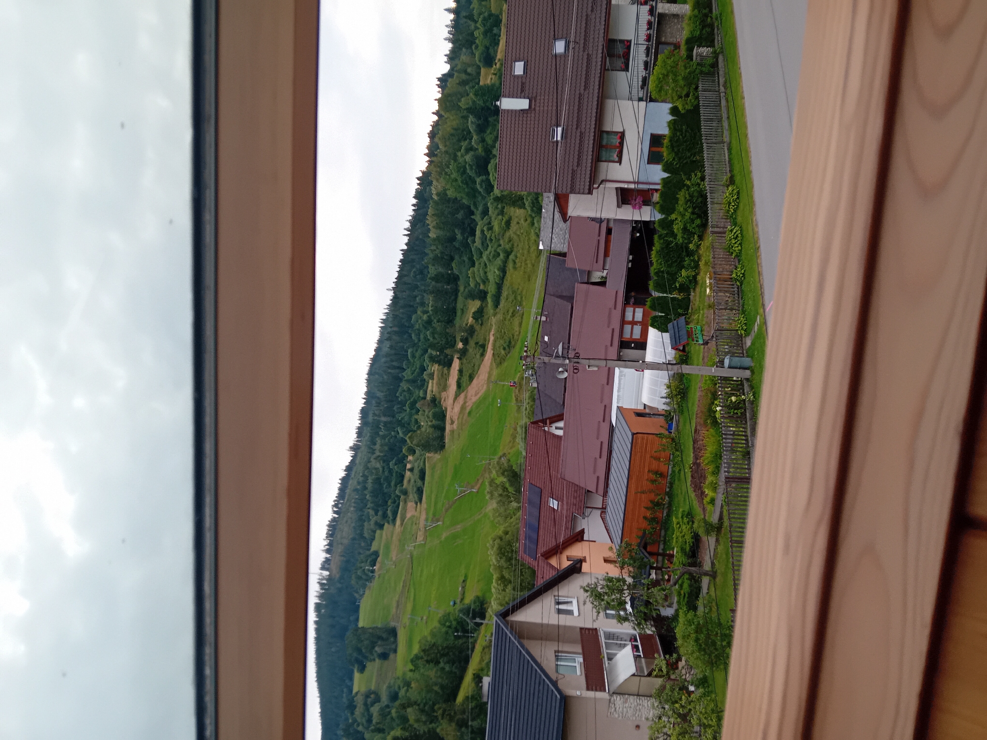 The image shows a view of a village in Zuberec, Slovakia, with a hill covered in trees and a ski slope in the background. The foreground features several houses with brown and red roofs, some with solar panels. A wooden fence runs along the edge of a property with a garden and a small wooden shed. A paved road is visible to the right of the houses, and utility poles are present. The sky is overcast, and the overall scene suggests a peaceful, rural setting. No people are visible in the image.