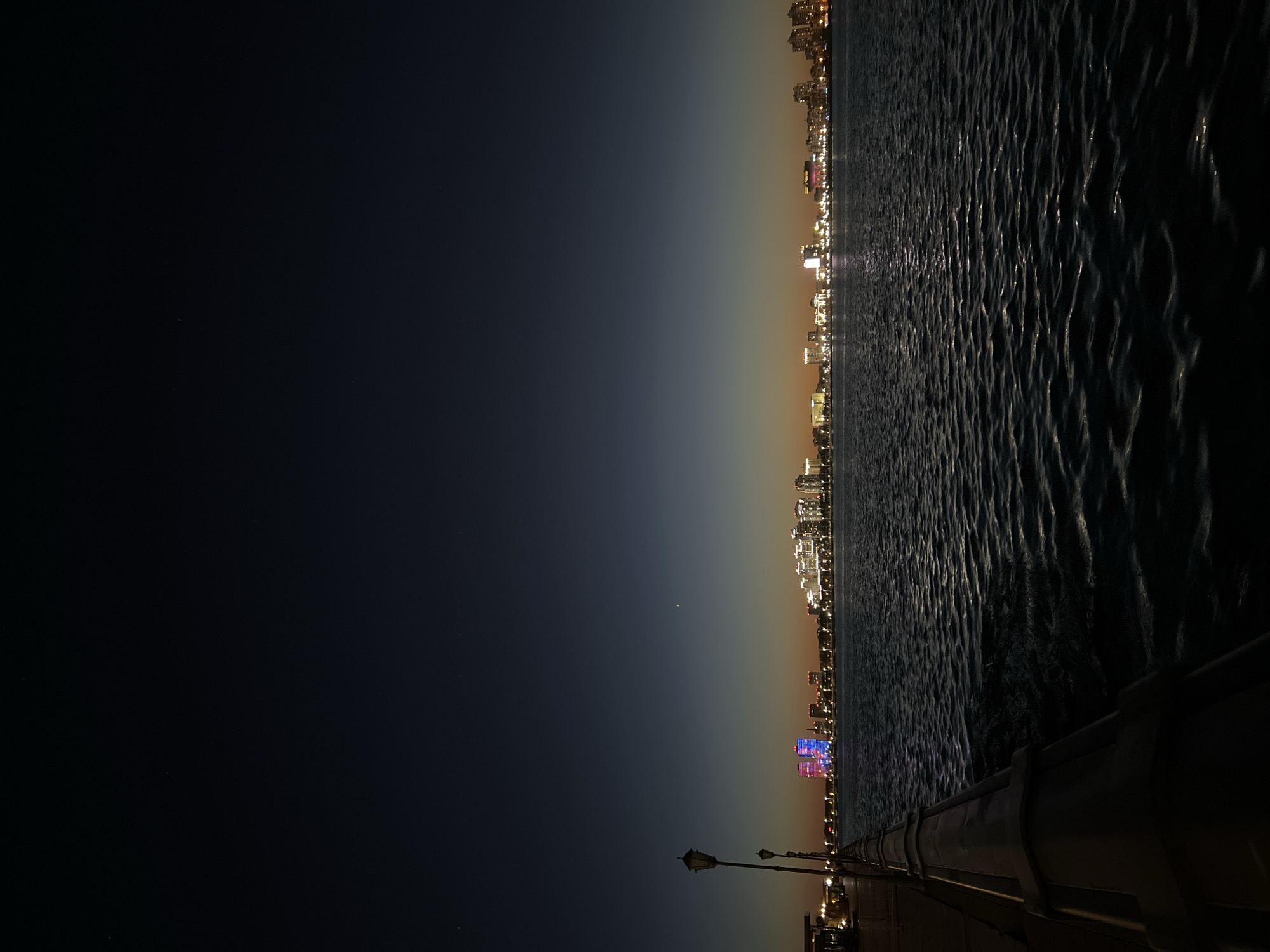 The image shows a night view of Kazan, Russia from a waterfront location. The right side of the image features dark, choppy water reflecting city lights. Across the water is the illuminated Kazan city skyline. Some taller buildings have distinct lighting displays. The left side of the image shows a dark, gradient-colored sky with a few visible stars. In the foreground, on the bottom right, there is a stone-like structure, possibly part of a boardwalk or pier. The dark silhouette of a lamppost is visible on the lower left.
