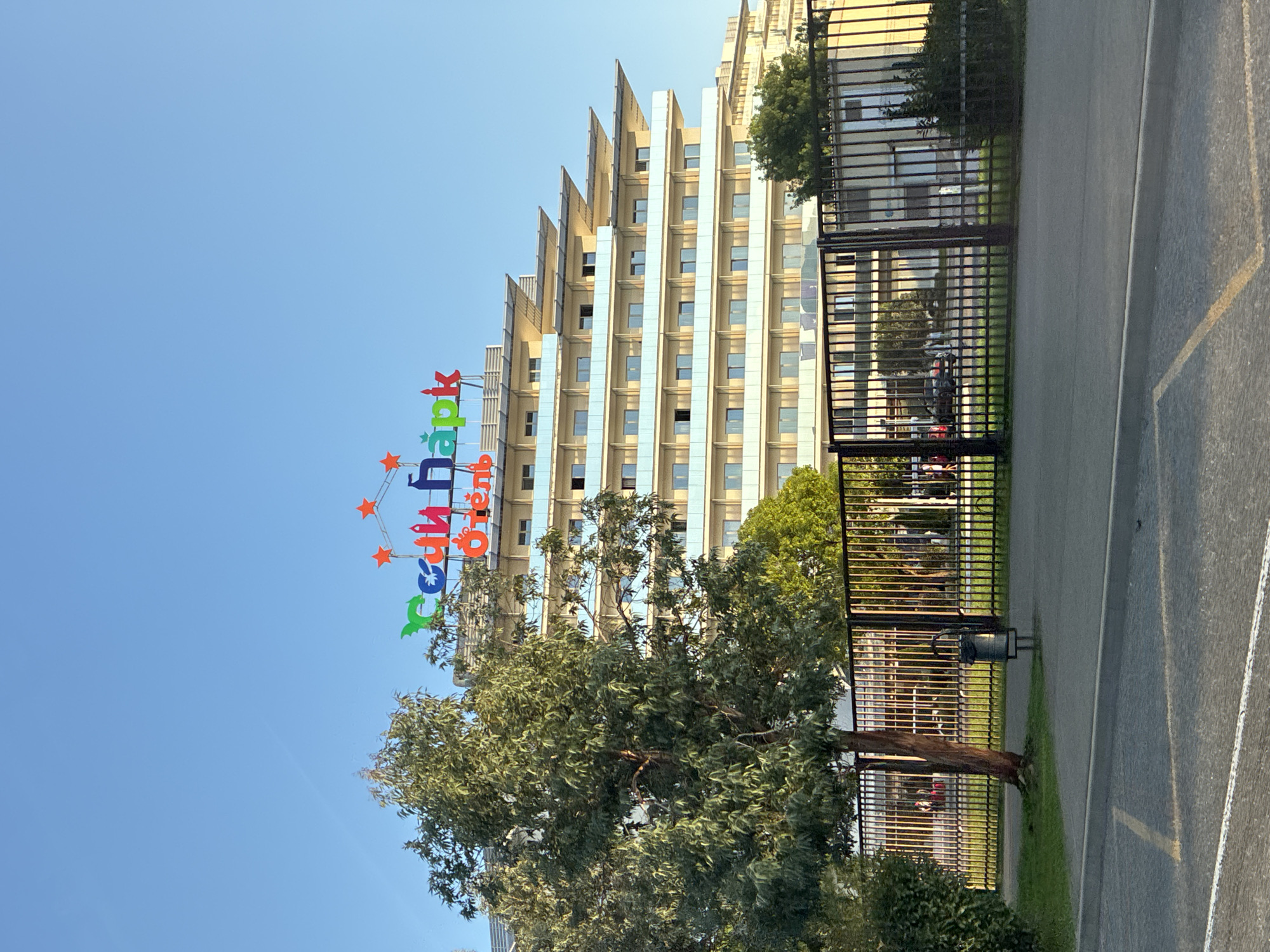 This image depicts the "Sochi Park Hotel" in Sirius, Russia, under a clear blue sky. The hotel building features a distinctive tiered design with multiple balconies and a brightly colored rooftop sign spelling out "Sochi Park" in Russian, accented with red stars and various figures. In the foreground, a black metal fence encloses the hotel grounds, with a lone tree and a trash can visible on a grassy area behind it. Beyond the fence, a paved area with visible road markings suggests a parking lot or street. The overall scene captures the facade of a hotel with an amusement park-themed branding, set against a backdrop of urban architecture and landscaping.