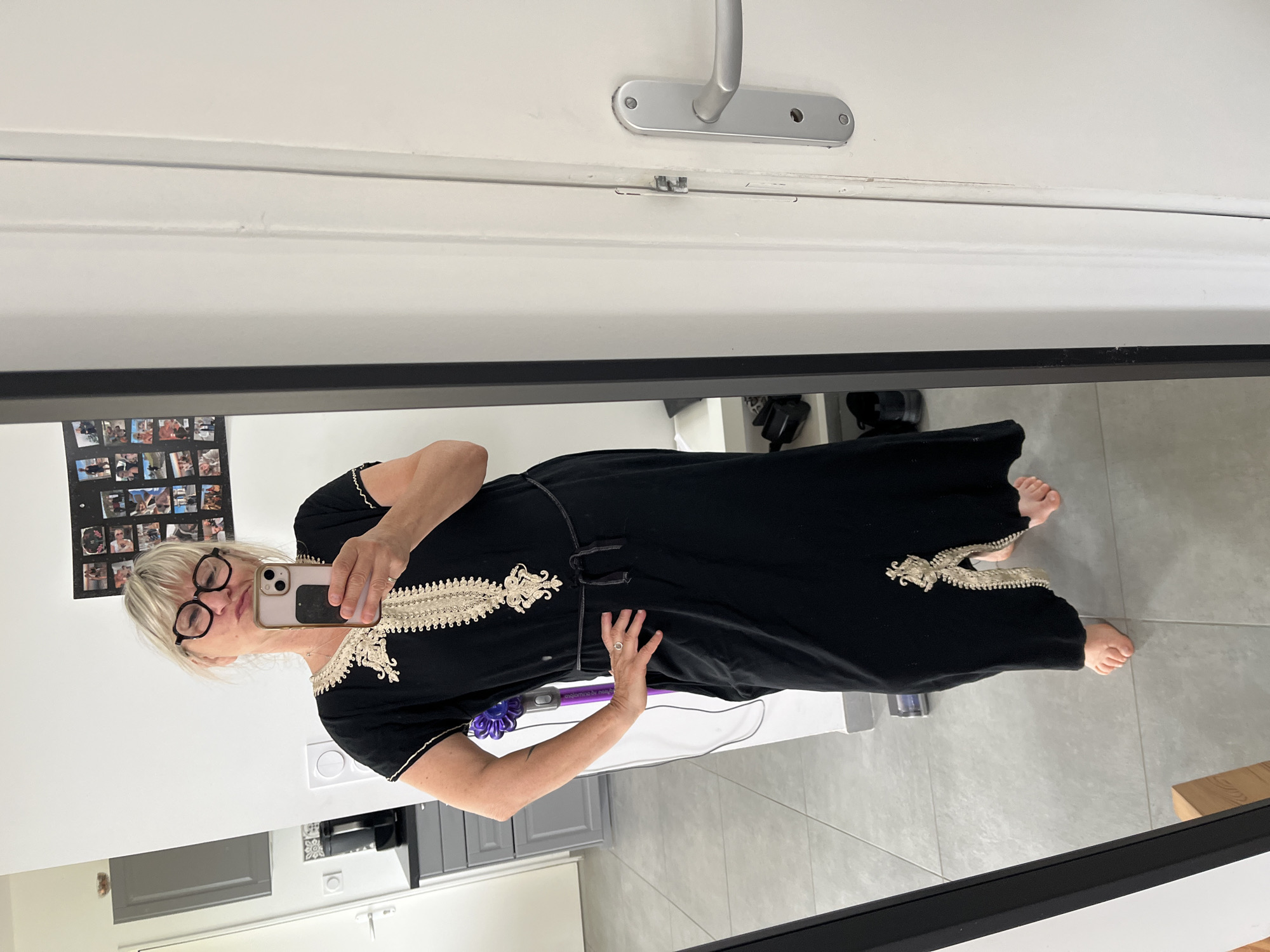 A woman is taking a mirror selfie indoors. She is wearing a black dress with light-colored embroidered trim and eyeglasses. She is holding a smartphone in her left hand and has a ring on her finger. The reflection reveals a room with light-colored walls and gray cabinets. A Dyson vacuum cleaner is visible in the background. The image does not provide any specific information to determine that the image was taken in the city of Chambœuf, France.