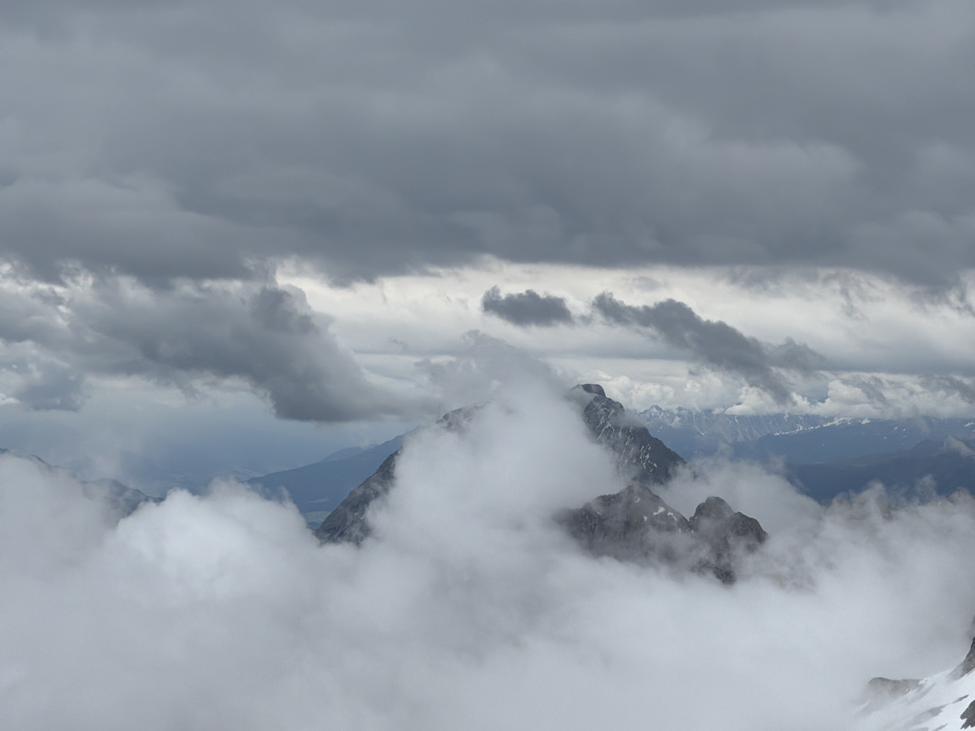 The image presents a mountainous landscape in Garmisch-Partenkirchen, Germany, under heavy cloud cover and extensive fog. The upper portion of the scene features a dense, dark gray overcast sky, with subtle variations in cloud density. Below this, thick white and light gray clouds and fog permeate the midground and foreground, partially obscuring the terrain. Several prominent, dark gray, rugged mountain peaks, composed of exposed rock, are visible emerging from and receding into these atmospheric conditions. Small patches of white, indicative of snow or ice, are present on some of the visible mountain surfaces, particularly towards the lower right. In the distant background, through breaks in the immediate cloud layers, faint, hazy blue mountain ranges can be discerned. There are no individuals, animals, or man-made structures present in the frame. The lighting is diffused and even, characteristic of a heavily overcast day.