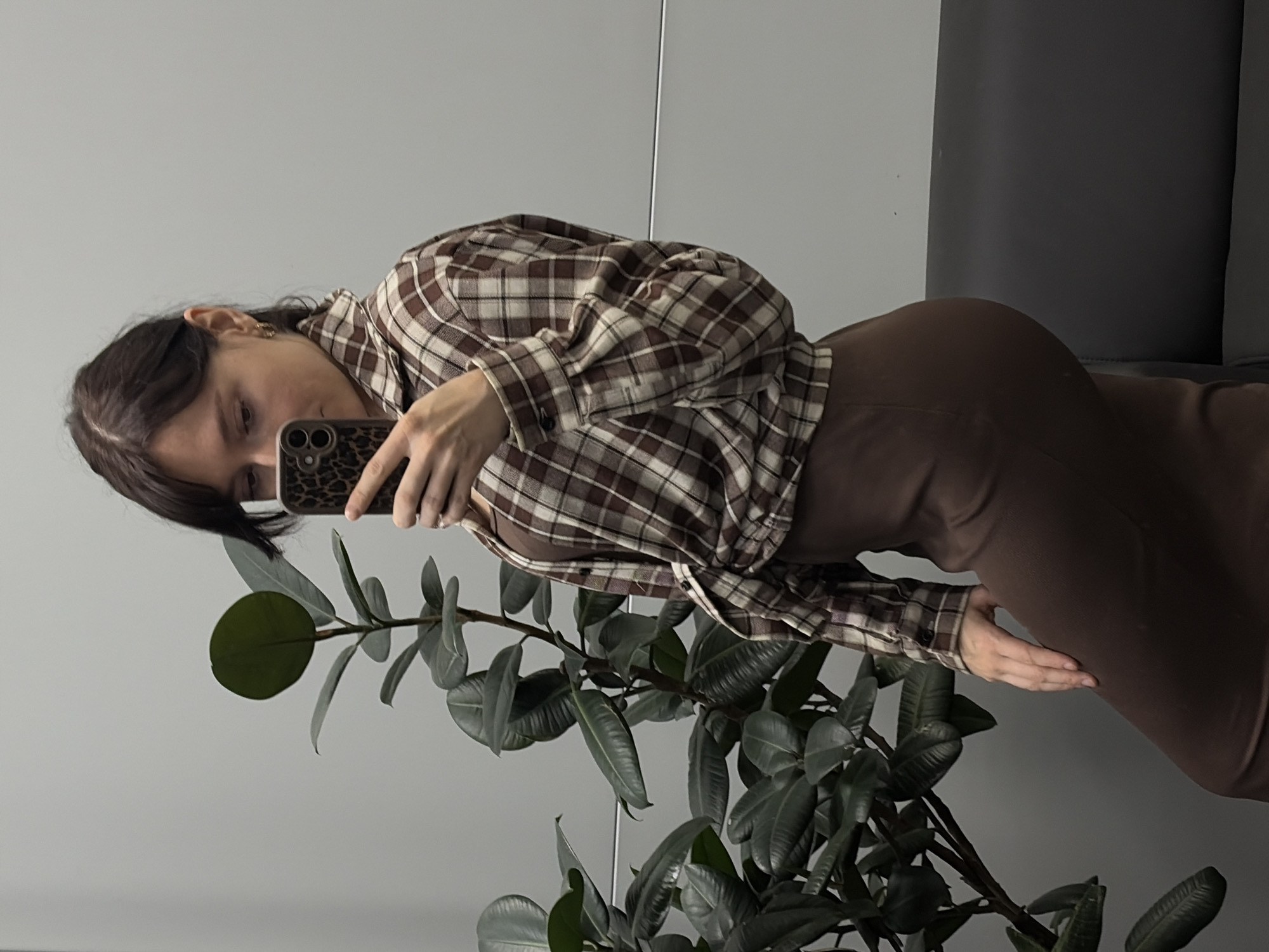 A young woman with dark hair and bangs, wearing a brown and beige plaid shirt tied at the waist and a brown fitted dress, is taking a mirror selfie in what appears to be an indoor space. She is holding a smartphone with a leopard-print case. To her left is a large Ficus elastica plant with dark green leaves. A dark gray sofa is visible to her right, and the background is a plain light gray wall. The scene is likely a home or office environment in Chaikovsky, Russia.