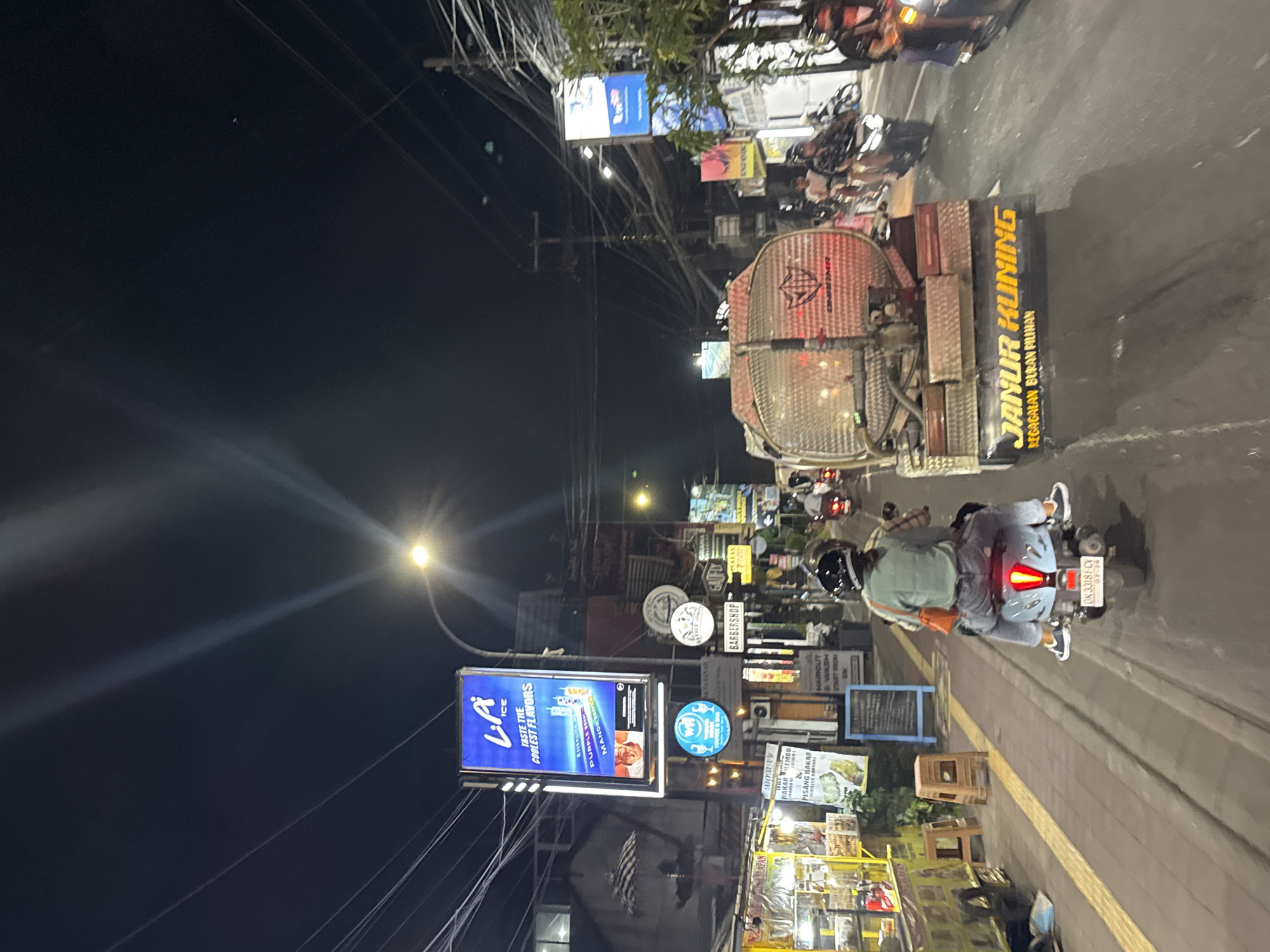 The image depicts a street scene at night in Kuta Selatan, Indonesia. A large truck with "JANUR KUNING" written on its side in yellow is present, alongside other vehicles, including a motorcycle being driven by a person wearing a helmet and a light-colored outfit. There are numerous signs advertising various businesses and products, including "LA Ice." Streetlights illuminate the scene, and overhead power lines are visible. A pedestrian sidewalk is on the right side of the image. The overall atmosphere suggests a bustling urban environment in the evening.