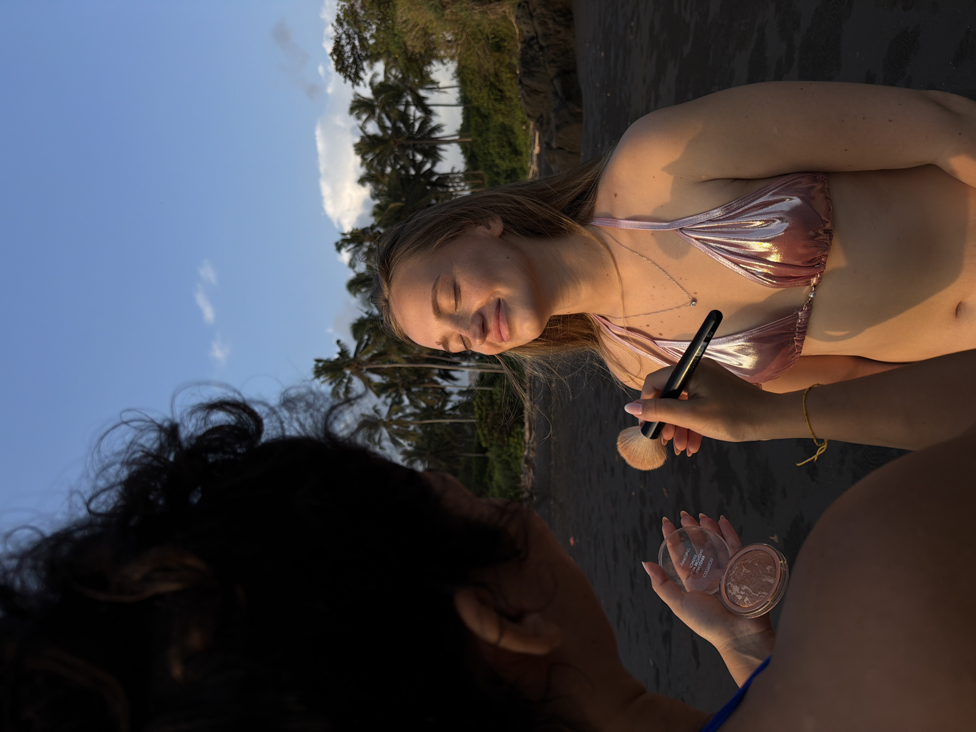 A young woman with blonde hair and freckles stands on a black sand beach, her eyes closed and a gentle smile on her face, as another person applies makeup to her cheek with a fluffy brush. The woman is wearing a shimmering pink bikini top with a delicate silver necklace. Behind her, palm trees line the horizon under a clear blue sky with scattered white clouds. The location is identified as Selemadeg Timur, Indonesia, with lush greenery and rocky outcrops visible in the background. The scene is captured during what appears to be sunset, with warm, golden light illuminating the subjects and the environment.