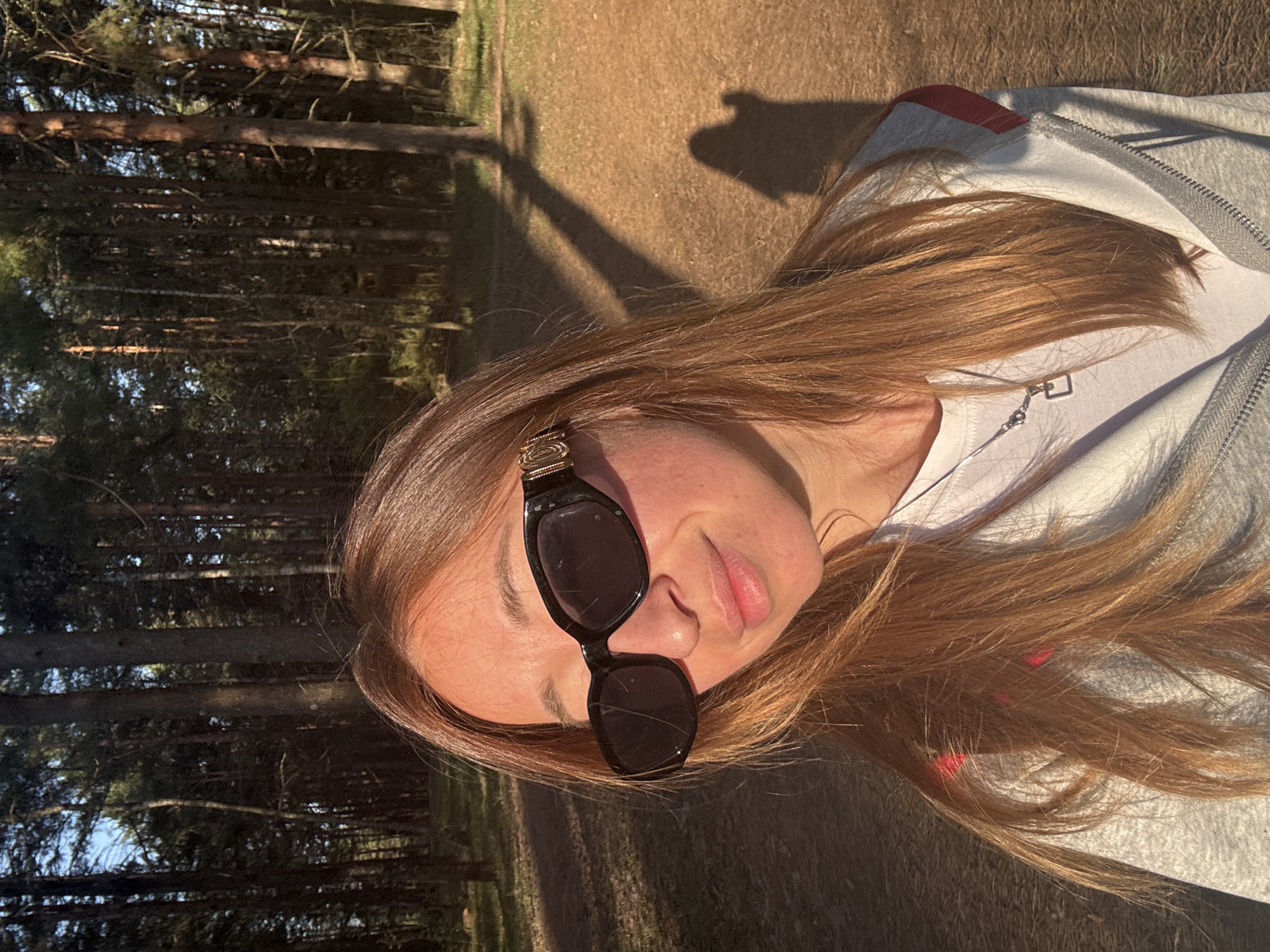 A photograph taken outdoors shows a woman with reddish-brown hair wearing black sunglasses and a white t-shirt under a grey zip-up hoodie with red accents. She is looking towards the camera with a slight smile. The background consists of a forest with tall, thin trees and sunlight filtering through the branches. The ground is covered in dry leaves or grass. The setting appears to be a wooded area, possibly a park or forest. There is no information to identify the specific location within Прикамский, Russia from the image.