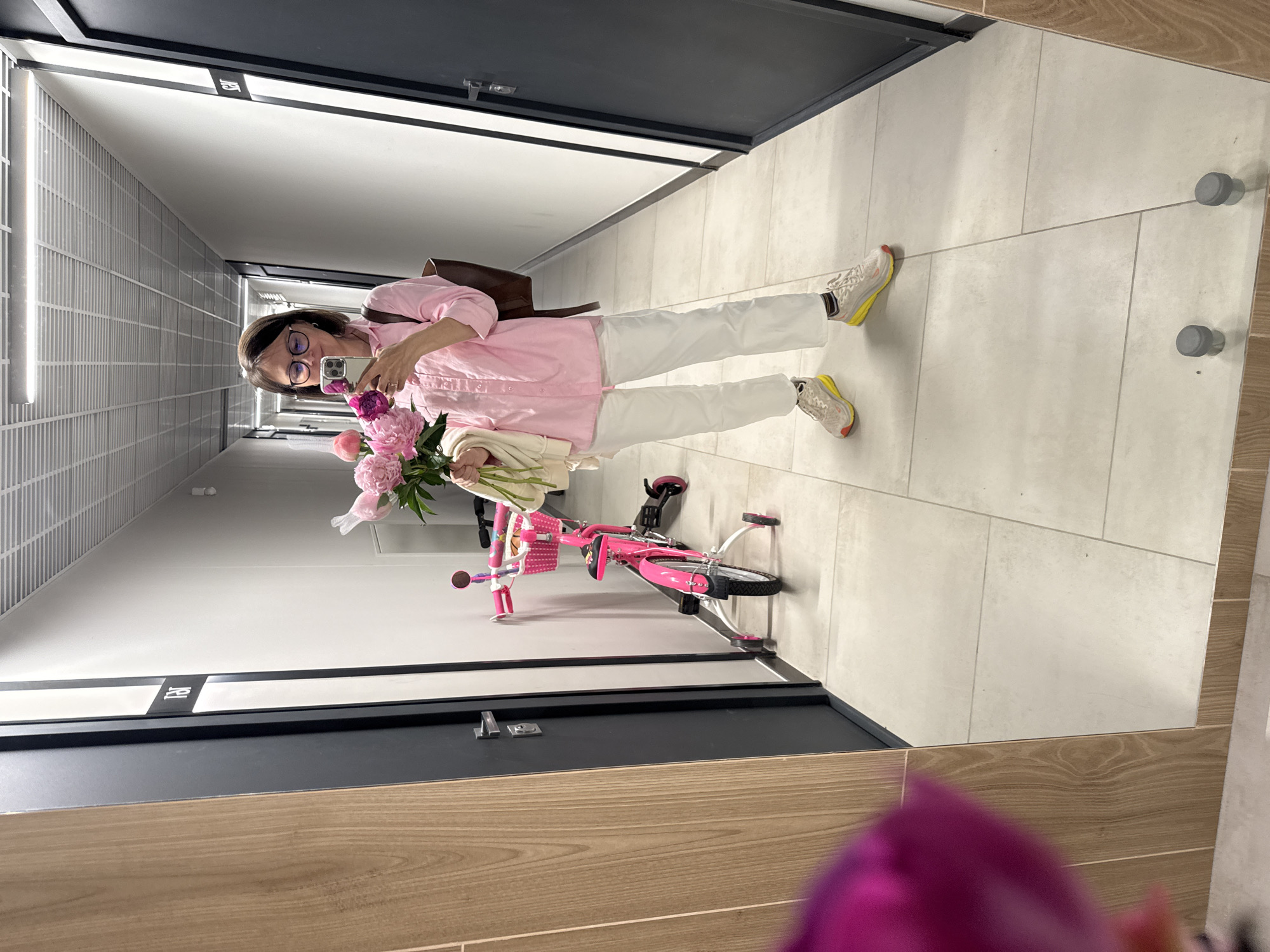 A woman stands in a well-lit, modern hallway and takes a mirror selfie. She is wearing a pink collared shirt, white pants, and athletic shoes. She holds a bouquet of pink flowers in her left hand and a phone in her right. To her left, a pink child's bicycle with training wheels is parked against the wall. The hallway features neutral-toned tiled flooring, white walls, and a metallic ceiling with fluorescent lighting. Two dark doors are visible on either side of the hallway, and a wooden-paneled section is on the left. The overall aesthetic suggests a residential or commercial building hallway in Izhevsk, Russia.