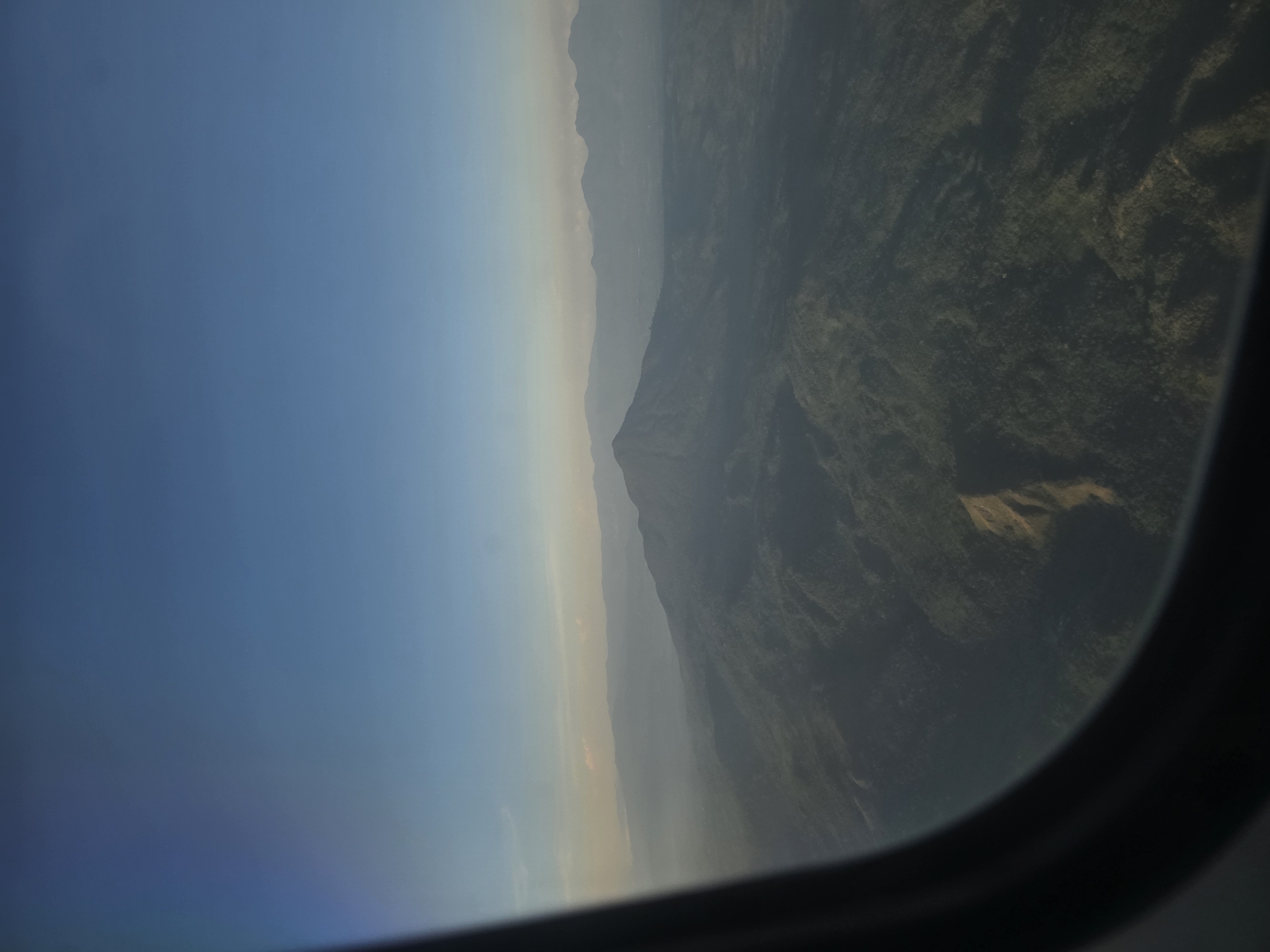 An aerial view captured through an airplane window shows a vast, verdant mountainous landscape under a clear blue sky. The foreground is dominated by the dark curve of the airplane window frame. The mountains, covered in dense forestation, stretch into the distance, becoming hazier and lighter in hue as they recede. In the middle ground, a prominent mountain peak stands out, its slopes also heavily forested. A distinct, lighter-colored scar or path is visible on one of the mountain slopes. The horizon is marked by a series of layered mountains and a faint haze that softens the distant view. The scene is devoid of any visible human presence or activity. The image does not provide any specific details that would allow for identification of the location as Ujongmesjid, Indonesia.