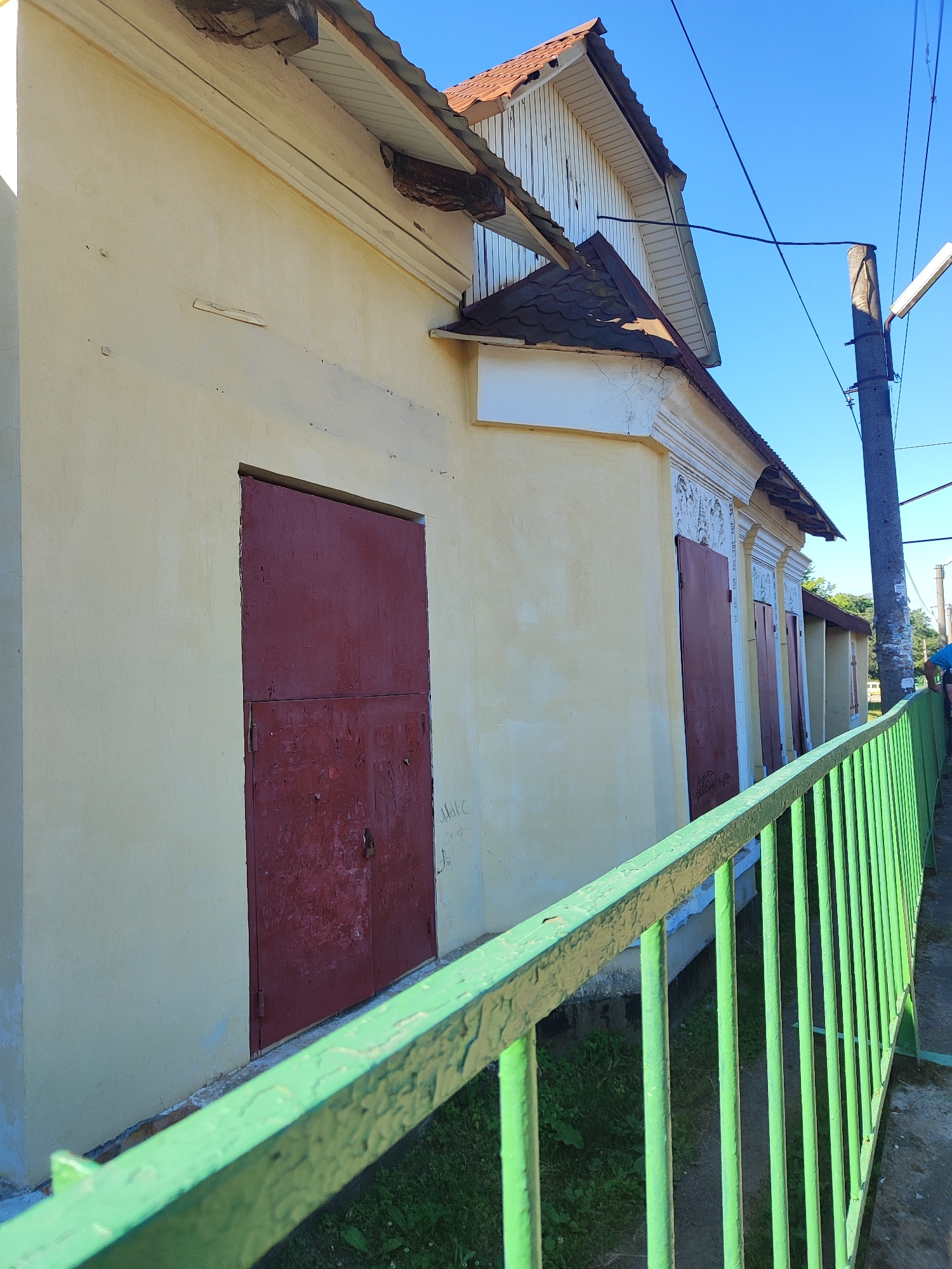 The image shows the exterior of a light yellow building with a red metal door on the left and several other red doors or shutters on the right. The building has decorative molding above the doors on the right. The roof is tiled with reddish-brown tiles, and there are exposed wooden beams under the eaves. A bright green metal fence runs along the foreground. In the background, there is a concrete utility pole with wires and a street light, and a clear blue sky. There is no visible activity or people. The location is described as Unknown, Unknown.