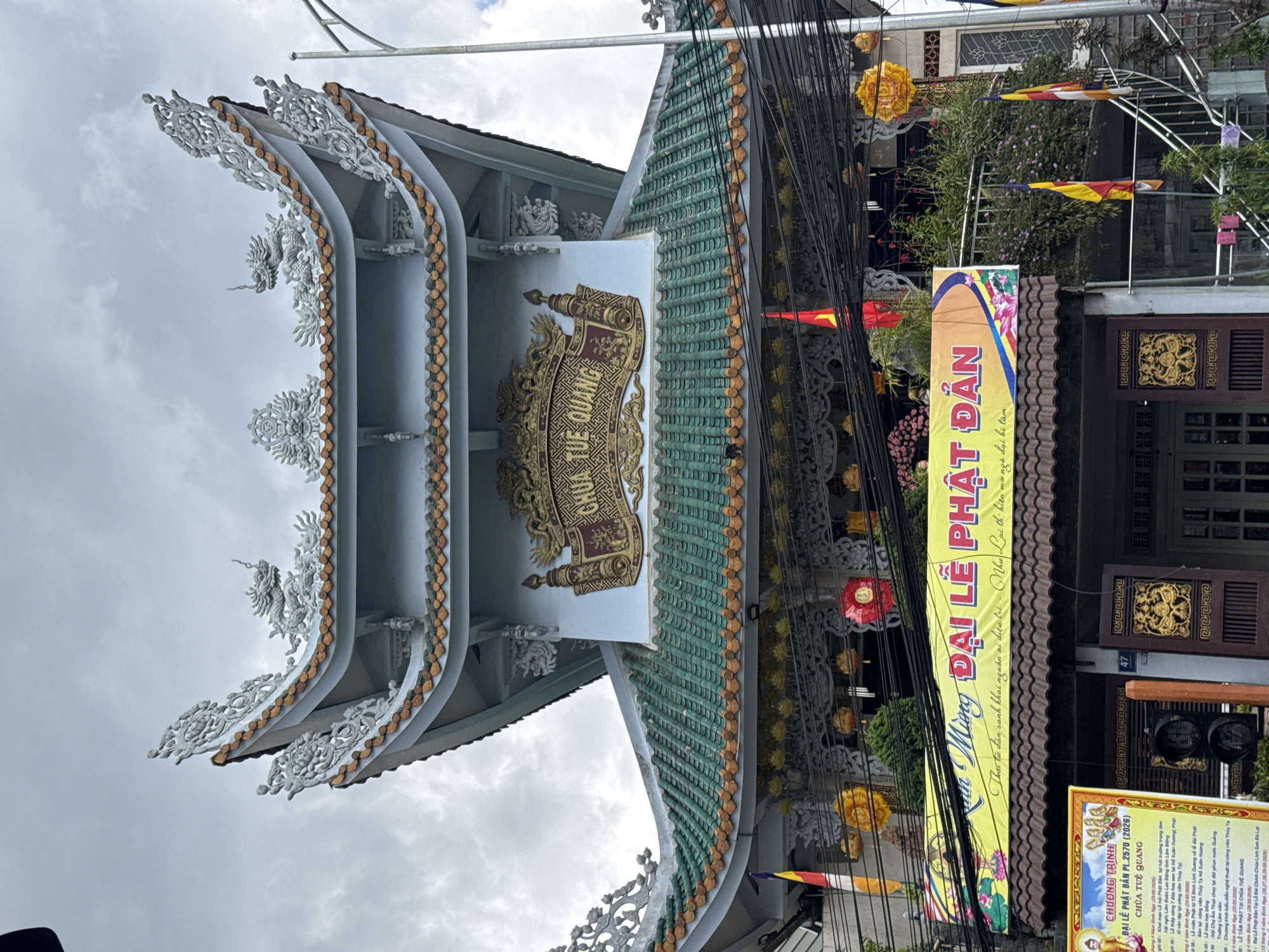 This is an eye-level shot of the ornate entrance of Chùa Tuệ Quang (Tuệ Quang Pagoda) in Cam Ly - Đà Lạt, Vietnam. The architectural details, including layered white eaves with dragon-themed ornaments and a sign in Vietnamese script, are central to the composition. A yellow banner advertising the celebration of the Buddha's Birthday (Phật Đản) is prominent on the right side. The overcast sky indicates the weather conditions. Utility wires cut across the upper part of the frame. Details on the sign and the building indicate preparations for religious events.