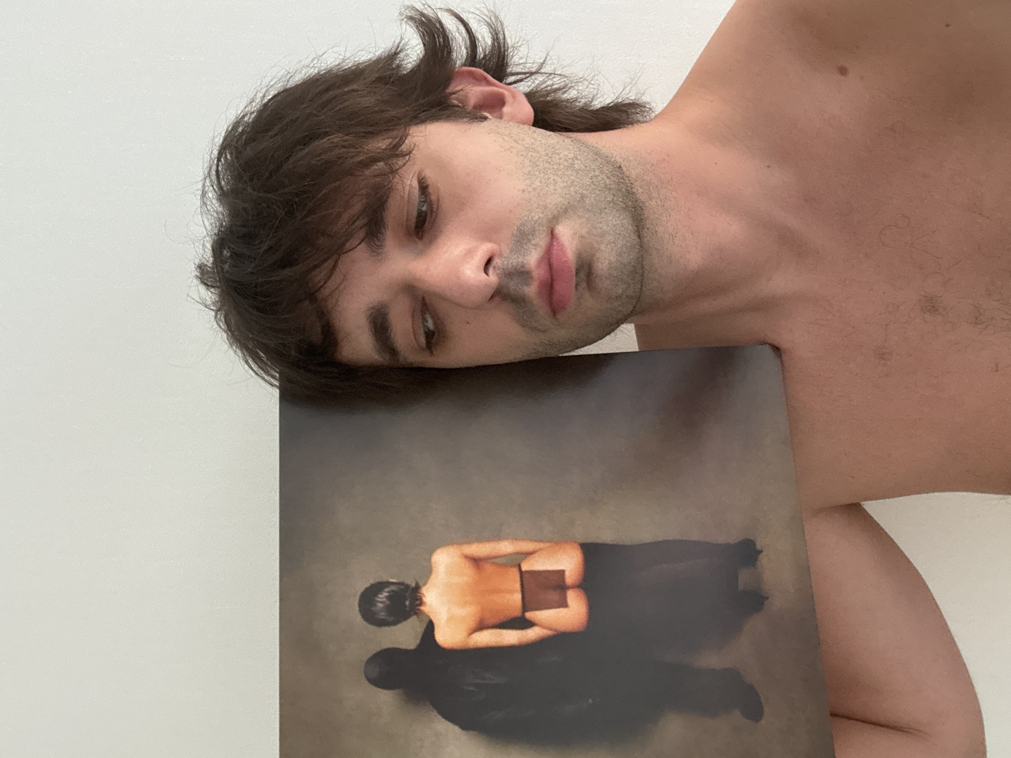 A shirtless person with dark, tousled hair and stubble is holding up a record album cover. The album cover depicts a person from behind, wearing minimal clothing, with a dark silhouette of another person behind them. The background of the image is a plain, light-colored wall. There is no indication of the location being Moscow, Russia.