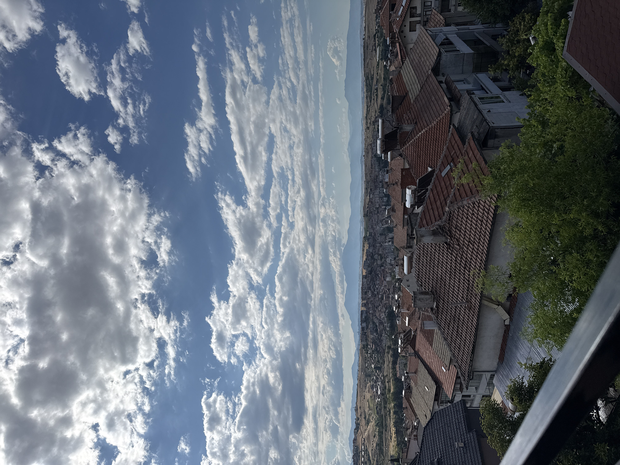 The image is a high-angle shot overlooking a residential area in Sandanski, Bulgaria. The foreground features a dark railing, partially obscuring the view of several houses with red-tiled roofs. Some houses have solar water heaters visible on their rooftops. Lush green trees are interspersed among the buildings.
In the midground, a denser collection of houses with predominantly red tile roofs fills the landscape. Beyond this, the town extends further, with buildings becoming less distinct as they recede into the distance. The background is dominated by rolling hills and a hazy mountain range under a bright blue sky with scattered cumulus clouds. The overall impression is a sunny day in a densely populated urban area adjacent to natural landscapes. There are no people or vehicles clearly visible in the image.