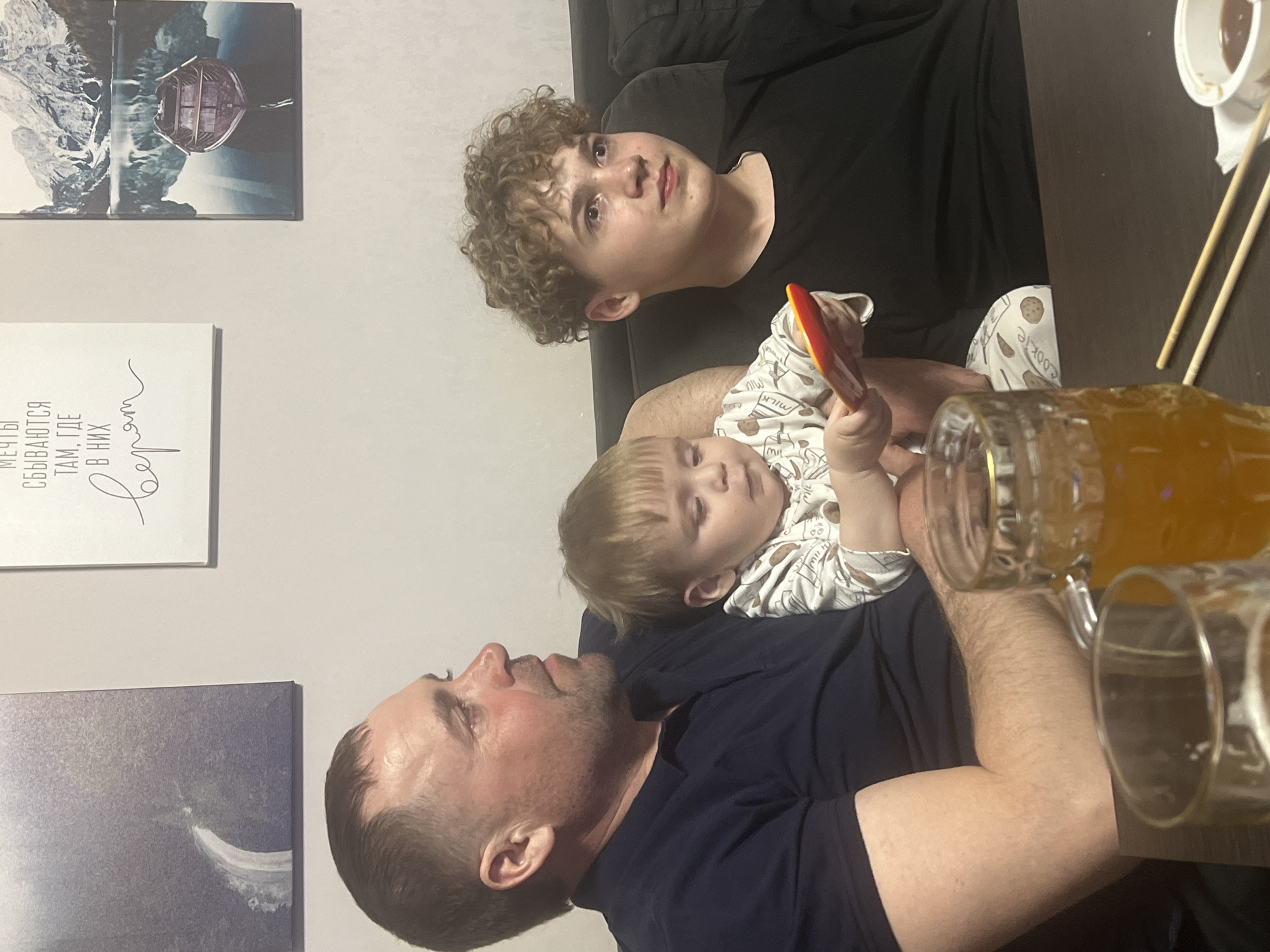 The image captures a casual indoor scene, likely in a home in Perm, Russia, featuring three males of different ages. The photograph is oriented vertically, showing the subjects and surroundings as if rotated clockwise by 90 degrees.
In the foreground, an adult man with short dark hair and a dark t-shirt lies on his back, looking upwards. A young child with blond hair and a white top adorned with cookie and milk patterns rests on his chest or shoulder, holding a red and yellow object (possibly a toy or piece of food). Both appear relaxed. To the top of the frame, an older boy or teenager with curly light brown hair is seated on a dark couch, looking with a neutral expression towards the camera's right.
On a dark wooden surface to the right, two glass mugs filled with an amber liquid (suggesting beer) are visible, along with a white bowl containing chopsticks and what appears to be a sauce or condiment. This arrangement hints at a casual meal or social gathering.
The background features light-colored walls adorned with several pieces of wall art. On the top left, a multi-panel artwork depicts a boat on water against a backdrop of mountains. Below it, a white canvas displays Russian text in a decorative script which reads: "МЕЧТЫ СБЫВАЮТСЯ ТАМ, ГДЕ В НИХ ВЕРЯТ," translating to "Dreams come true where they are believed in." A third, more abstract blue and white artwork is visible on the bottom left. The overall atmosphere is intimate and relaxed, suggesting an evening spent indoors.