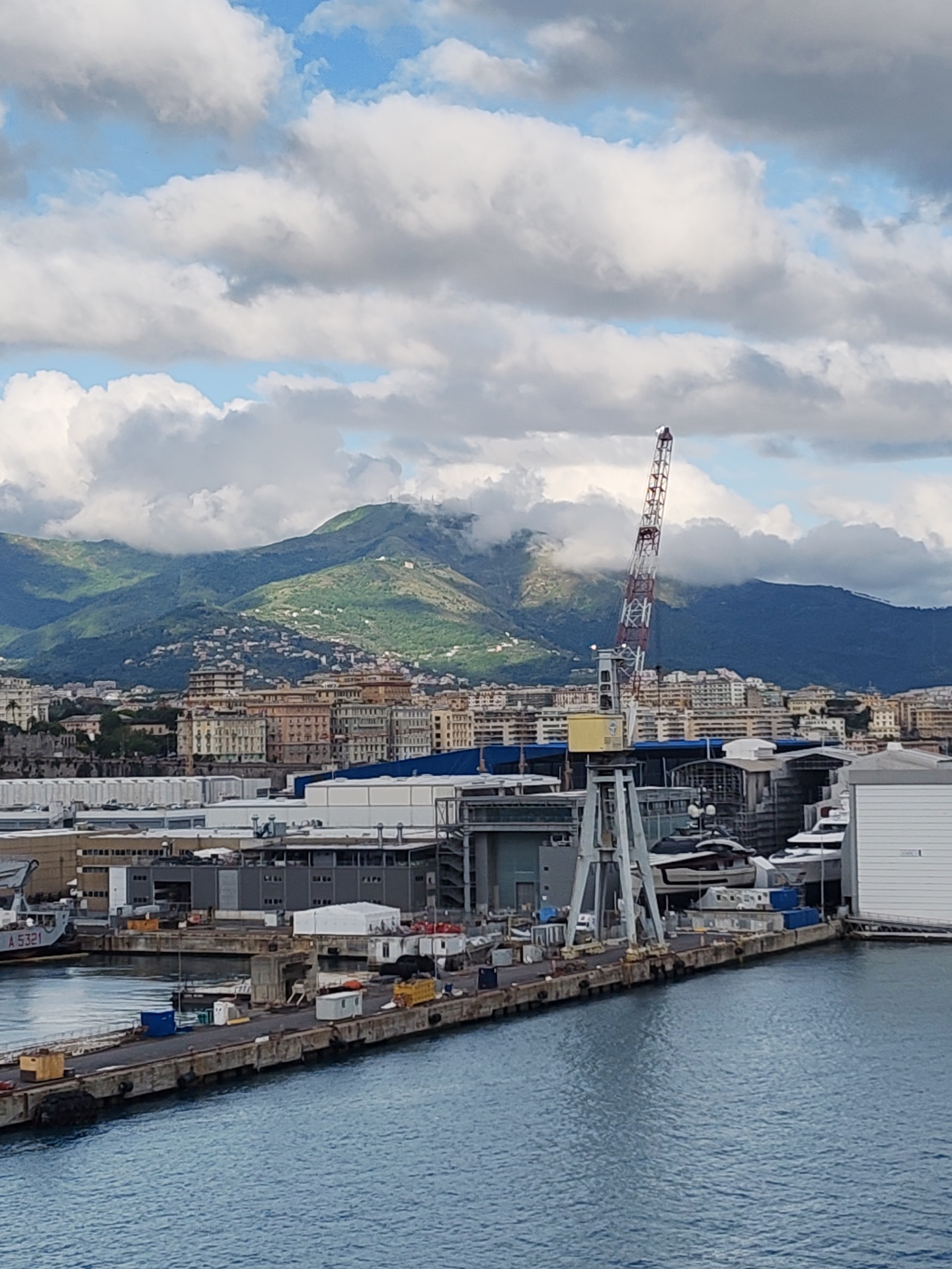 The image shows a port scene in Genoa, Italy. In the foreground is a concrete pier extending into the water, with various containers and small structures along its length. The water is calm, reflecting the sky. Behind the pier are industrial buildings and cranes, suggesting a shipyard or cargo port. A large crane with a red and white lattice boom is prominent. The cityscape of Genoa rises in the middle ground, with a mix of residential and commercial buildings. In the background, mountains are partially obscured by clouds. The sky is blue with numerous fluffy white clouds.