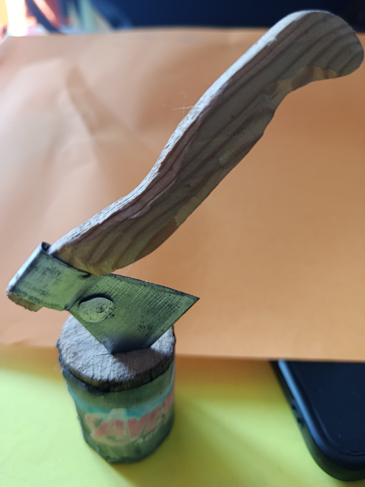 An extreme close-up shot focuses on a miniature, handmade axe. The axe features a crude wooden handle with visible grain and indentations, attached to a metal blade. The blade is rectangular with one sharpened edge, attached to the handle by a metal cap. The axe head is embedded into a small, cylindrical base, possibly a piece of wood or a rolled-up material, decorated with colorful, illegible markings. The axe is positioned at an angle, with the blade facing downwards. The background is out of focus, showing a transition from a soft orange surface to a bright yellow surface. A dark object, possibly a smartphone, is partially visible in the lower right corner. The image does not contain any elements that would allow for identification of its location in Biysk, Russia, or any other specific geographic area.