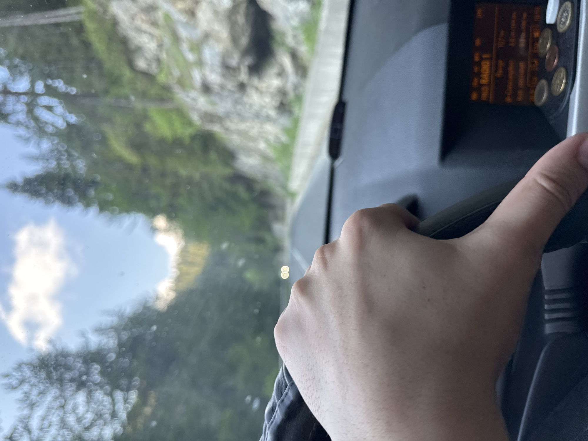 A right hand, light-toned with fine hair, grips a black steering wheel on the left side of the frame. The car's dashboard, composed of gray and black plastic, is visible below the hand. To the right of the steering wheel, a digital display, possibly a car radio, shows blurry orange text, including "RND RADIO 1". Below the display, several metallic coins are arranged on a dark, textured surface. Through the windshield, an asphalt road extends forward, marked by white lane lines. A concrete barrier or guardrail lines the left side of the road. Beyond the road, dense coniferous trees and rocky outcrops characterize a mountainous environment. A steep, rocky incline with sparse vegetation is visible on the right side. The sky above is blue with scattered white clouds, indicating daytime conditions. The scene depicts a vehicle in motion on a mountain road, specifically located in Sokolovtsi, Bulgaria.