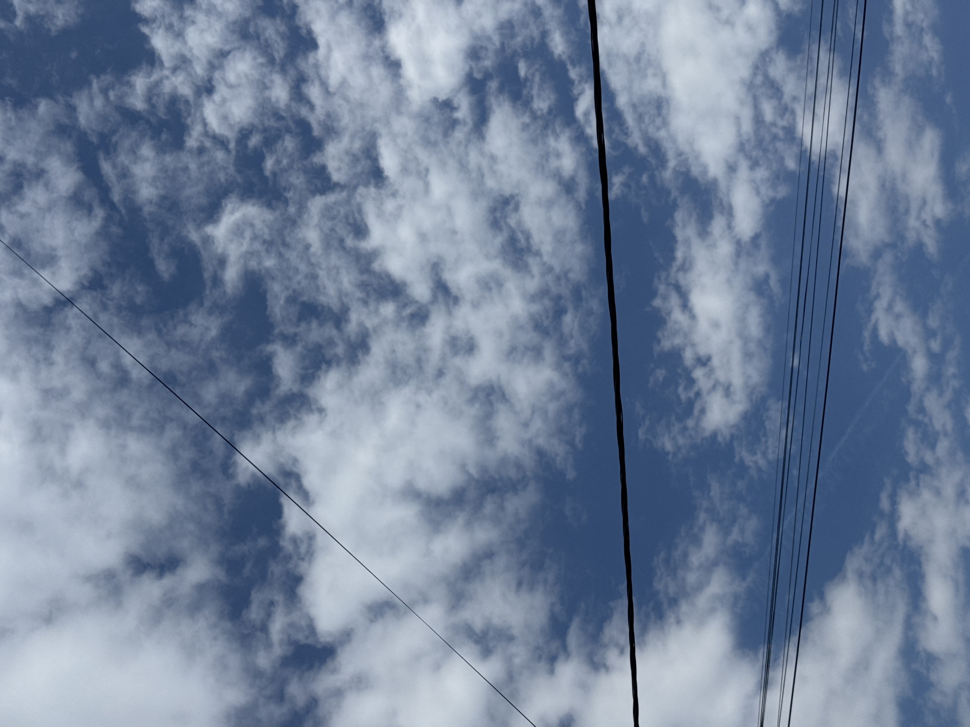 The image shows a view of the sky in Šušanj, Montenegro, filled with patchy, white clouds set against a blue sky. Several black power lines run across the image from different directions. Some are singular, while others are grouped together. The scene is quiet and focused on the natural and built environment.