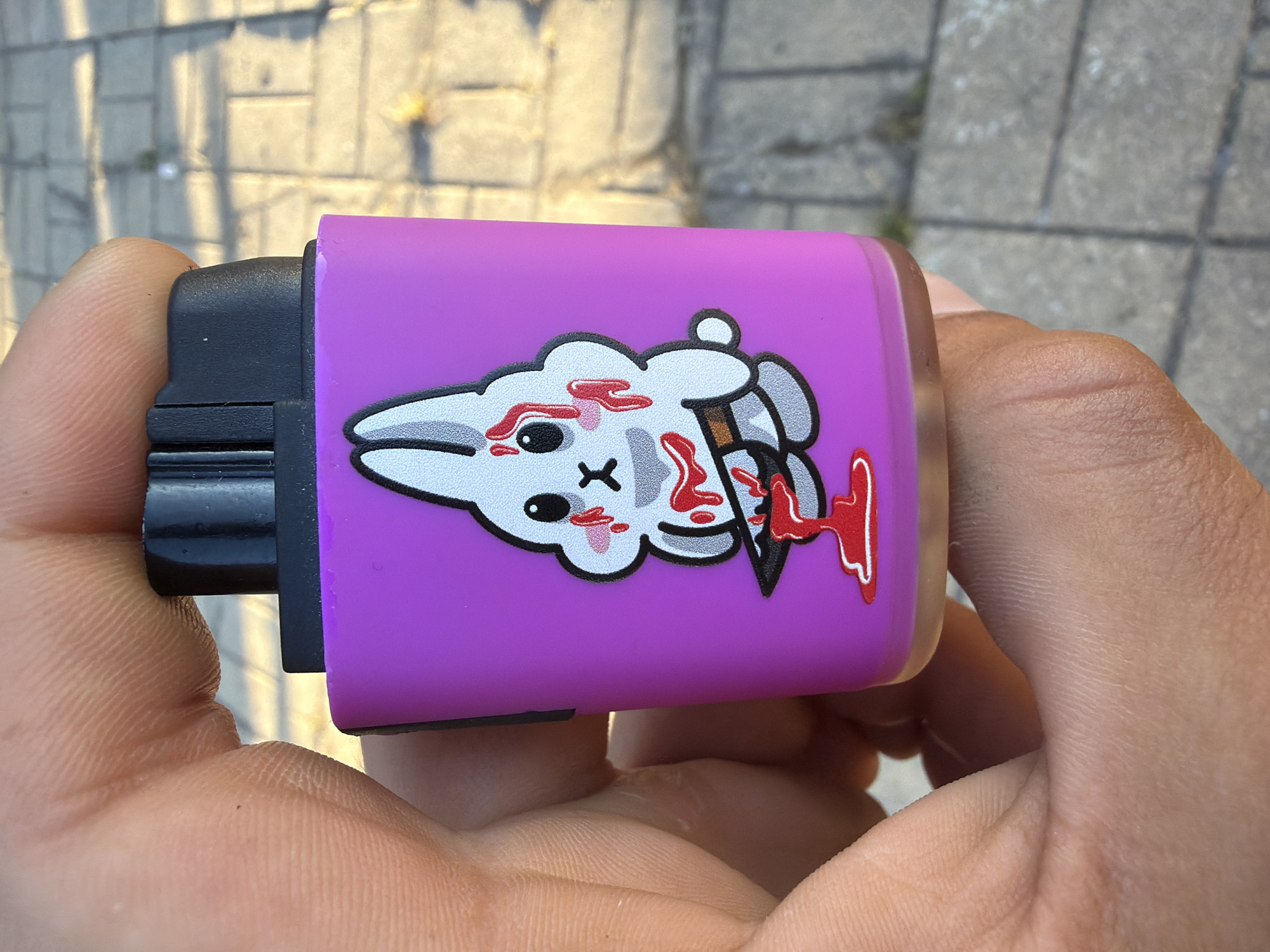 A close-up shot shows a person's hand holding a bright purple device, possibly a vape pen or a lighter, with a cartoon image of a bunny. The bunny is depicted with large, sad eyes, blushing cheeks, and is covered in what appears to be blood. The bunny is also holding a knife, with more blood splattered around it, suggesting a violent or dark theme. The background is out of focus, showing a paved surface with a brick-like pattern and some green vegetation, indicating an outdoor setting. The lighting suggests it is daytime, with sunlight casting shadows.