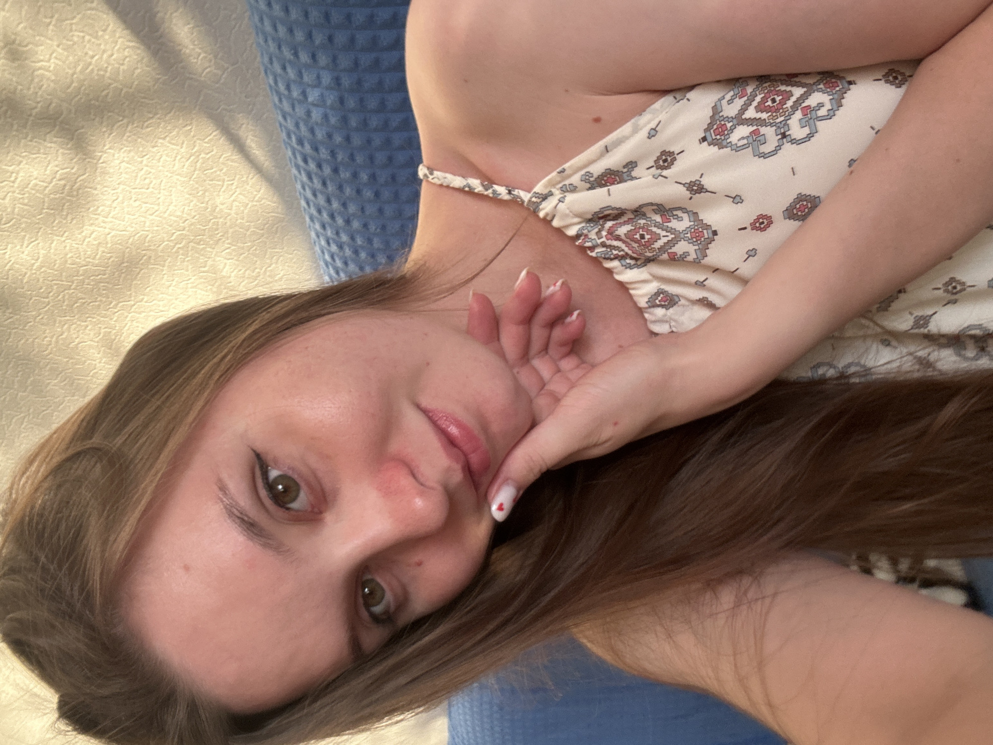 The media file shows a close-up, vertically oriented, portrait-style photograph of a young woman lying down. She is facing the camera and appears to be resting on a textured blue surface and a beige textured surface. Her head is tilted, and her right hand is gently touching her face, near her cheek and mouth. Her left arm is bent and her hand is also visible. She has long, brown hair that is spread out around her. The woman is wearing a light-colored, sleeveless top with a patterned design. The pattern is geometric, featuring diamond shapes and other small motifs in colors like red, blue, and beige.
Her facial expression is relaxed, and she is looking directly at the camera. Her eyes are open and appear to be brown. She has light-colored makeup on, including eyeliner and mascara. Her lips are slightly parted and appear to be glossy. Her fingernails are painted white with a small red heart on one of them. The lighting suggests it might be daytime, possibly indoors or outdoors in soft light, as there are subtle shadows cast on the beige surface. The overall mood is casual and relaxed. There is no visible text in the image. The provided location context is Noviy, Russia, which might be the setting for this photograph.