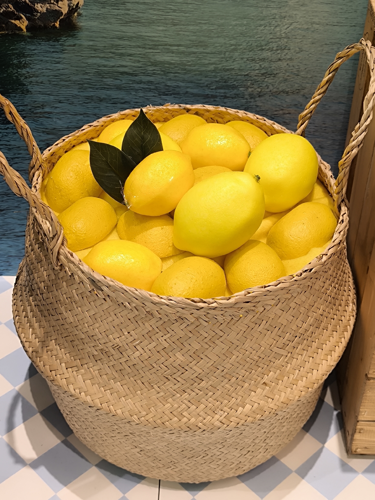 A medium shot depicts a woven basket filled to the brim with bright yellow lemons, two dark green leaves nestled amongst the fruit. The basket is positioned on a blue and white checkered surface. The background features a muted image of water. The location is Riga, Latvia.