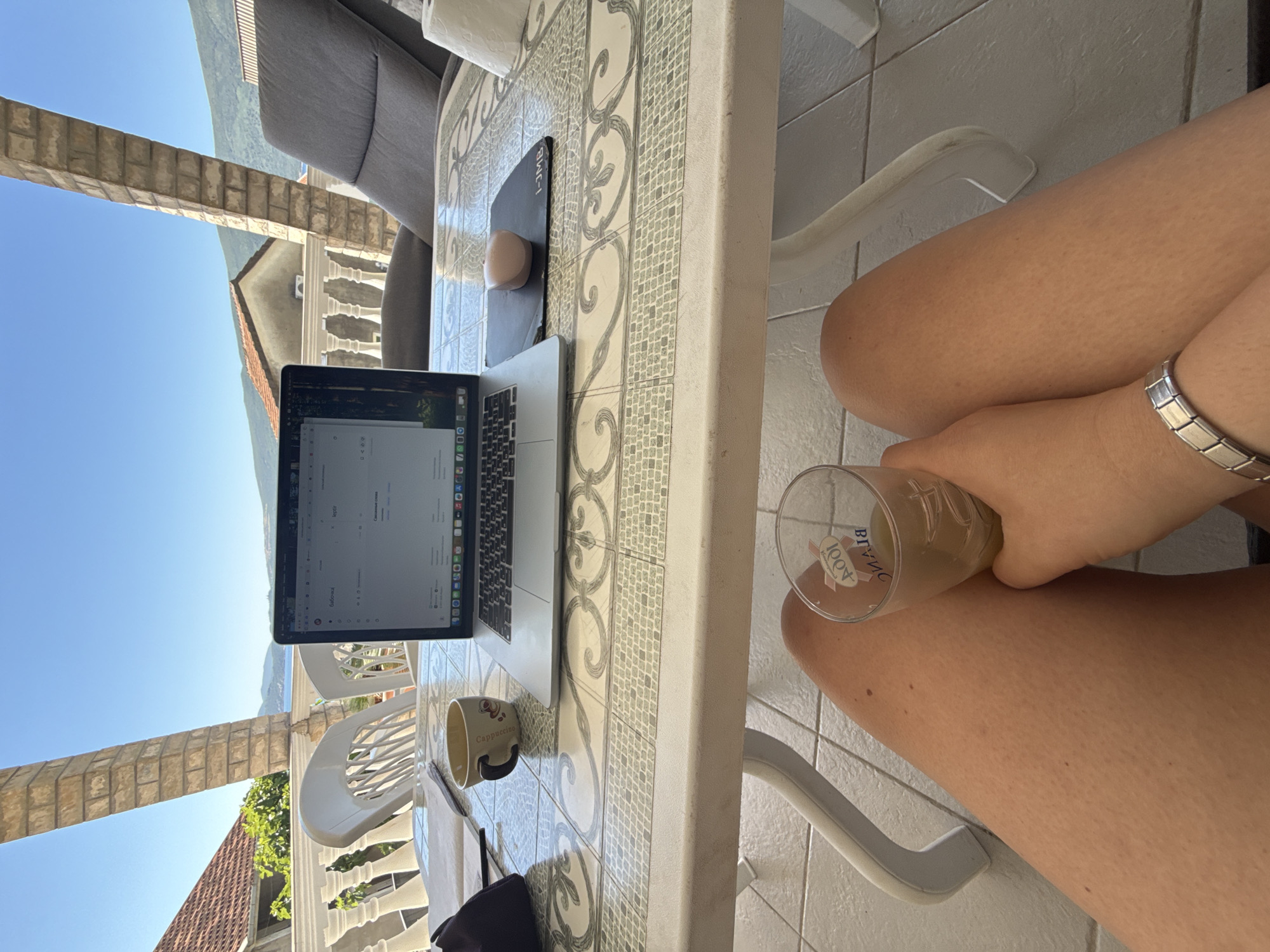 The image captures a person working on a laptop at a table outdoors in Bijela, Montenegro. The person is seated at a white, tiled table with a laptop, mouse, mousepad, and a mug labeled "Cappuccino." They are holding a glass with a beverage. The person's leg and wrist, adorned with a silver bracelet, are visible. In the background, there is a view of buildings, a balcony with balustrades, a glimpse of the sea, and mountains under a clear blue sky. A partial view of outdoor furniture is also present. The image suggests a work-from-home or vacation setting.
